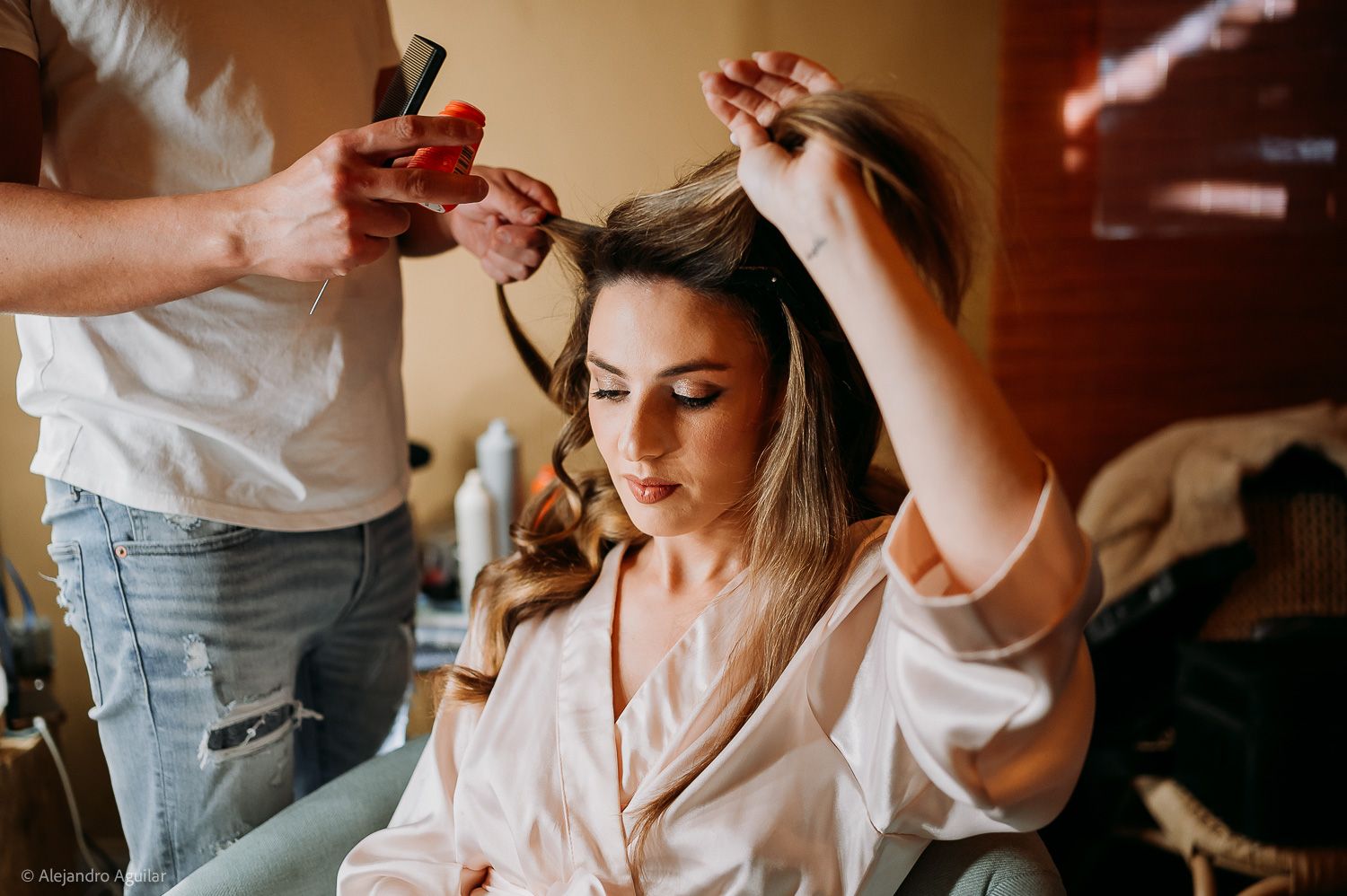 A bride sits while a hairstylist curls her hair during wedding preparations in a warmly lit room.