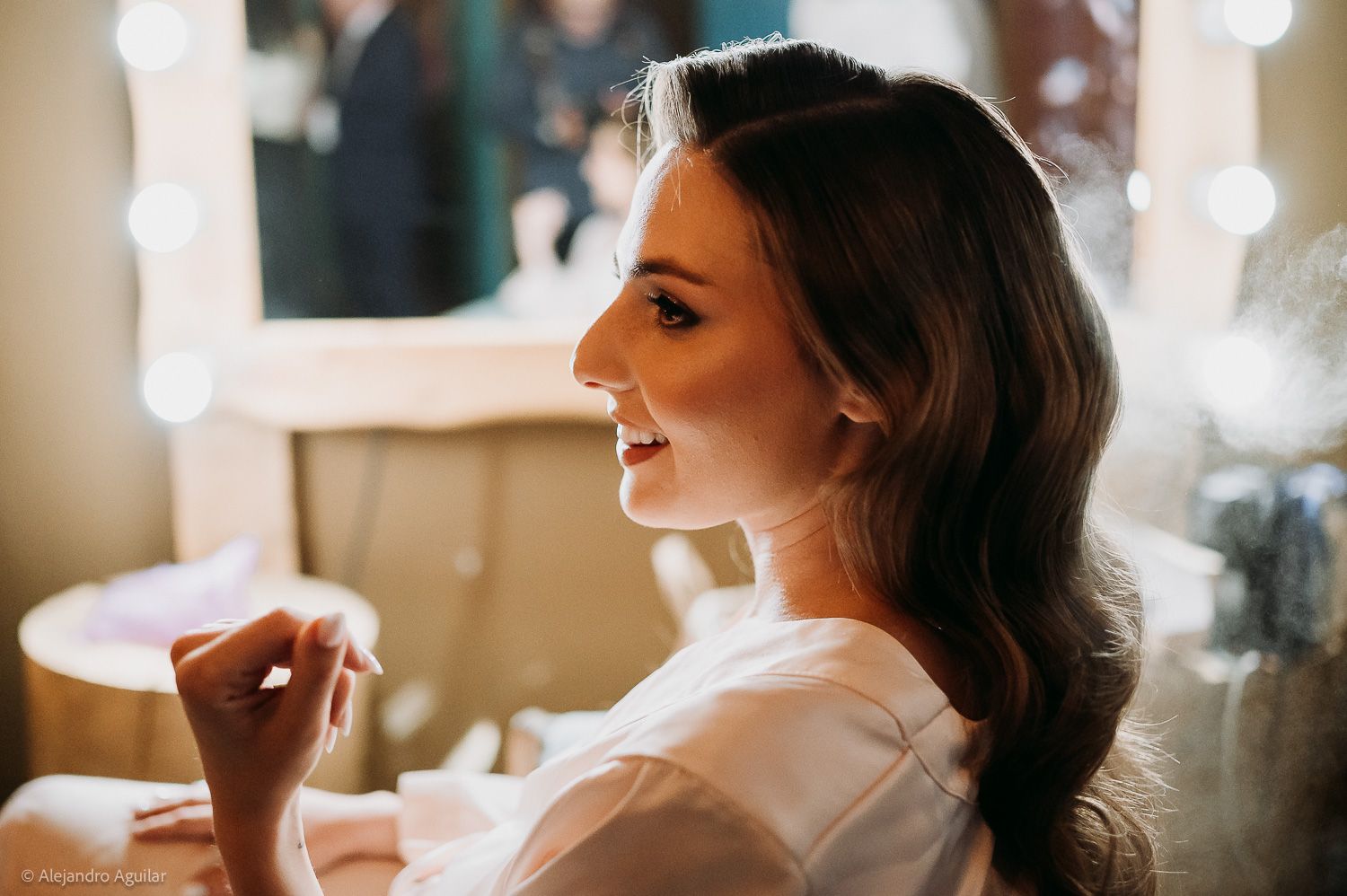 Smiling bride in white robe applying perfume while getting ready, with soft bokeh lights in background.