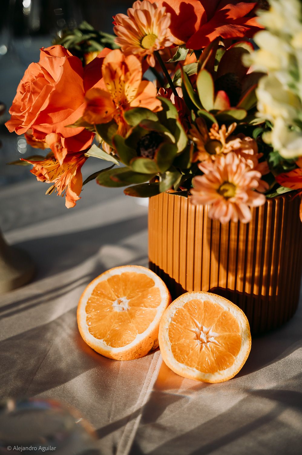 Halved orange in front of an orange ribbed vase with vibrant orange flowers and greenery on a linen cloth.