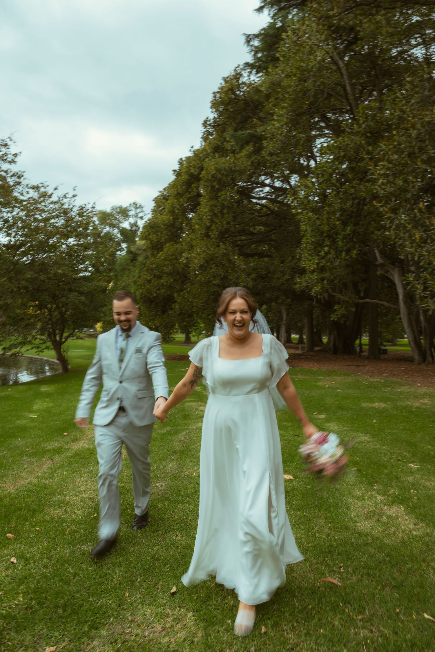 A joyful bride in a white gown and groom in a gray suit walk hand-in-hand through a lush green park.