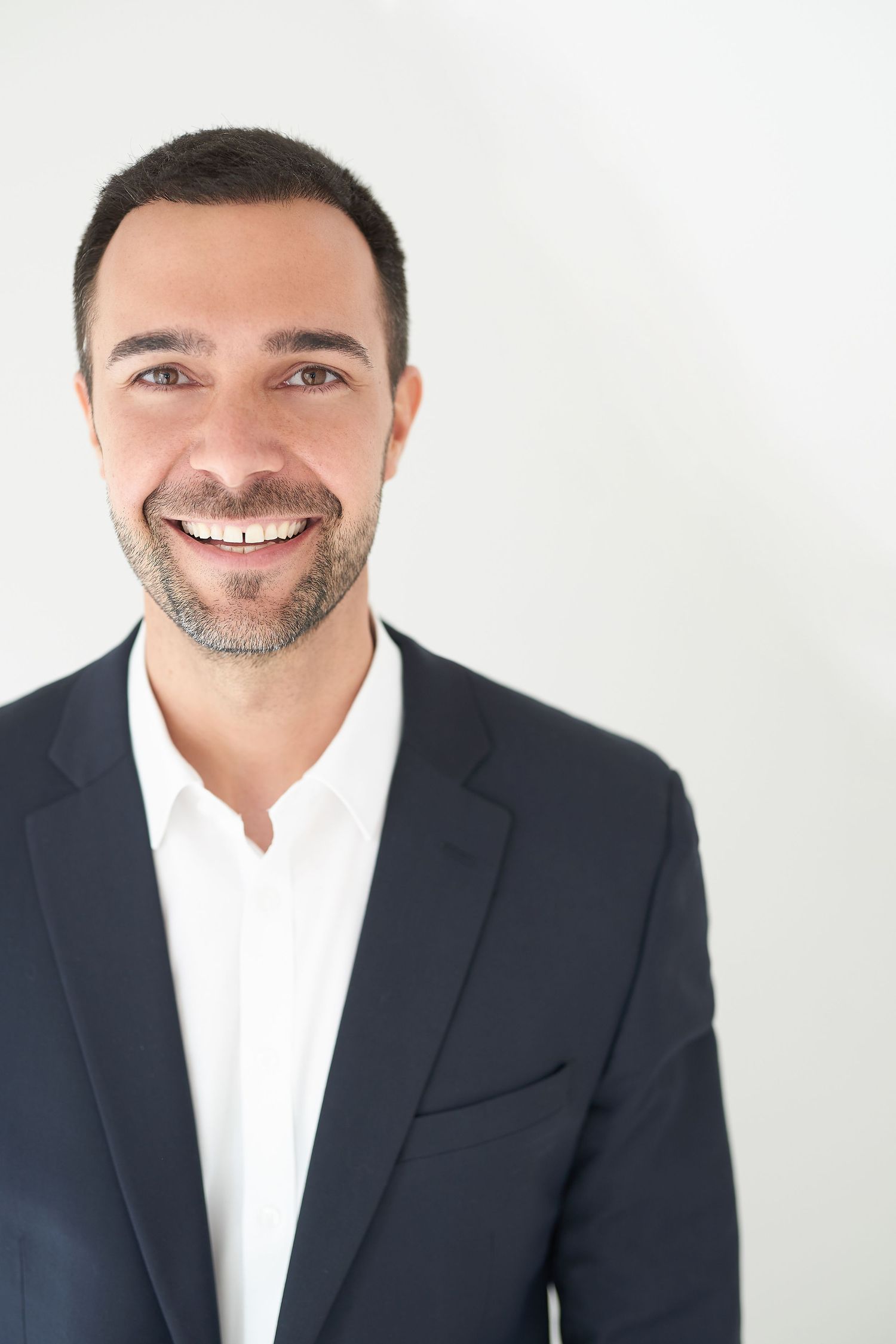 Manuguerra Fotografie - Lächelnder Geschäftsmann in dunklem Blazer und weißem Hemd posiert selbstbewusst vor einem hellen Hintergrund.