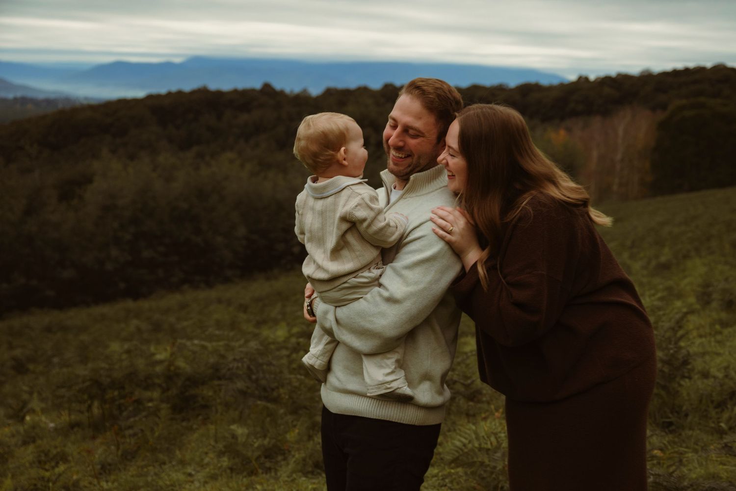 A happy family of three enjoys an outdoor moment, with parents smiling at their toddler against a scenic green hillside.