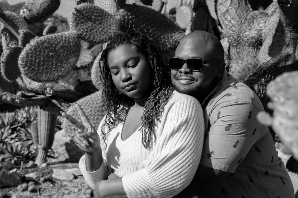 Black and white photo of a couple embracing in front of a large cactus garden backdrop.