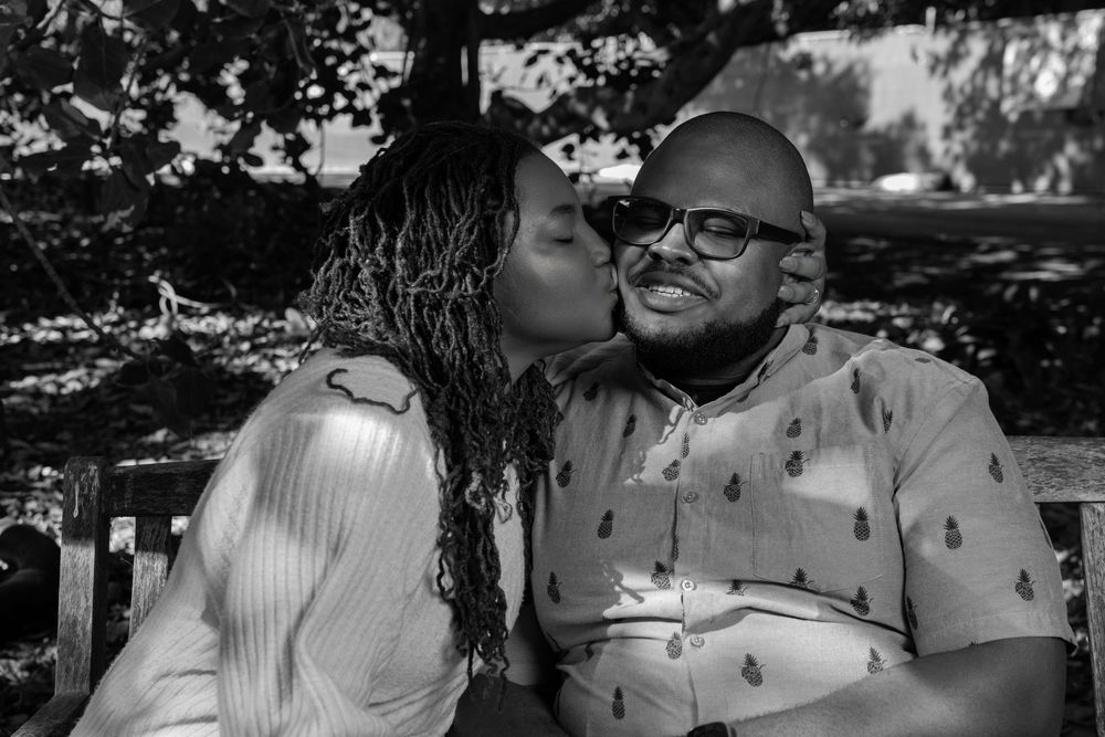 Black and white photo of a woman kissing a smiling bald man on the cheek while sitting on a bench outdoors.