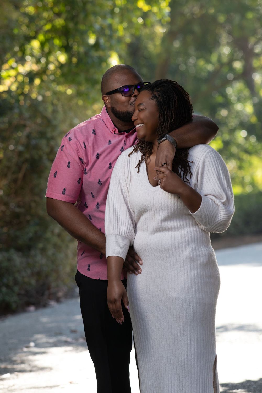 A man in a pink shirt kisses a smiling woman in a white dress outdoors among green trees.