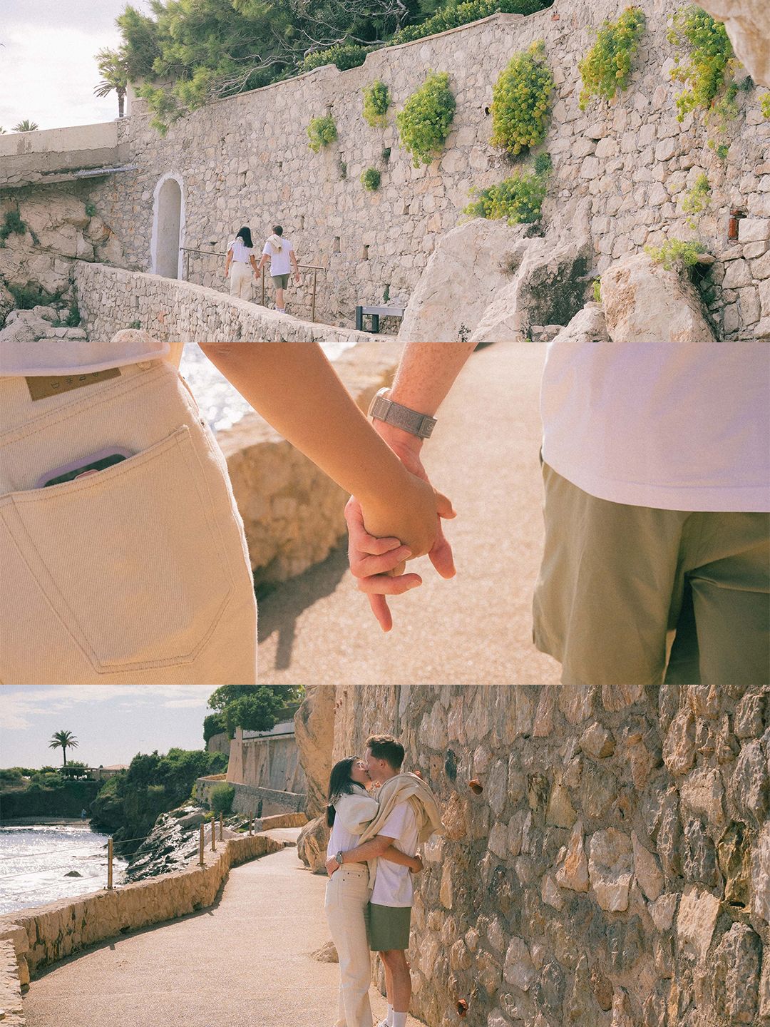 Couple holding hands and kissing near a sunlit stone wall along a scenic coastal walkway.