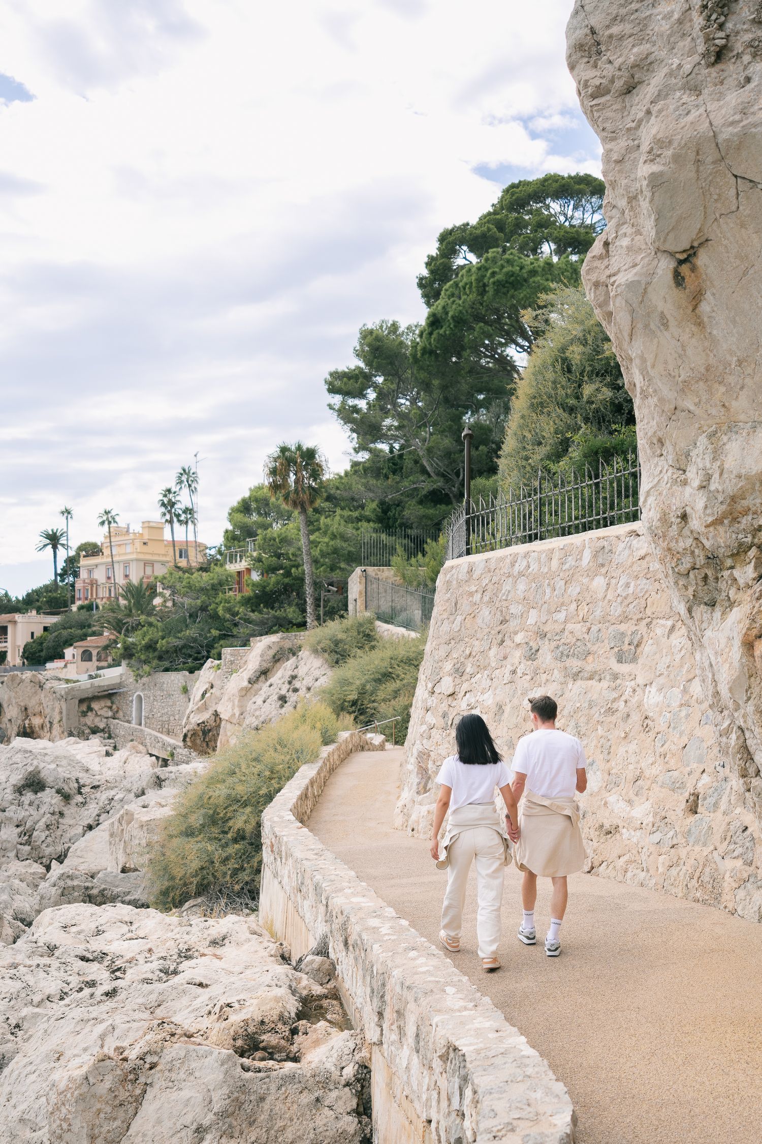 Couple walking hand in hand along scenic stone coastal pathway surrounded by lush greenery.