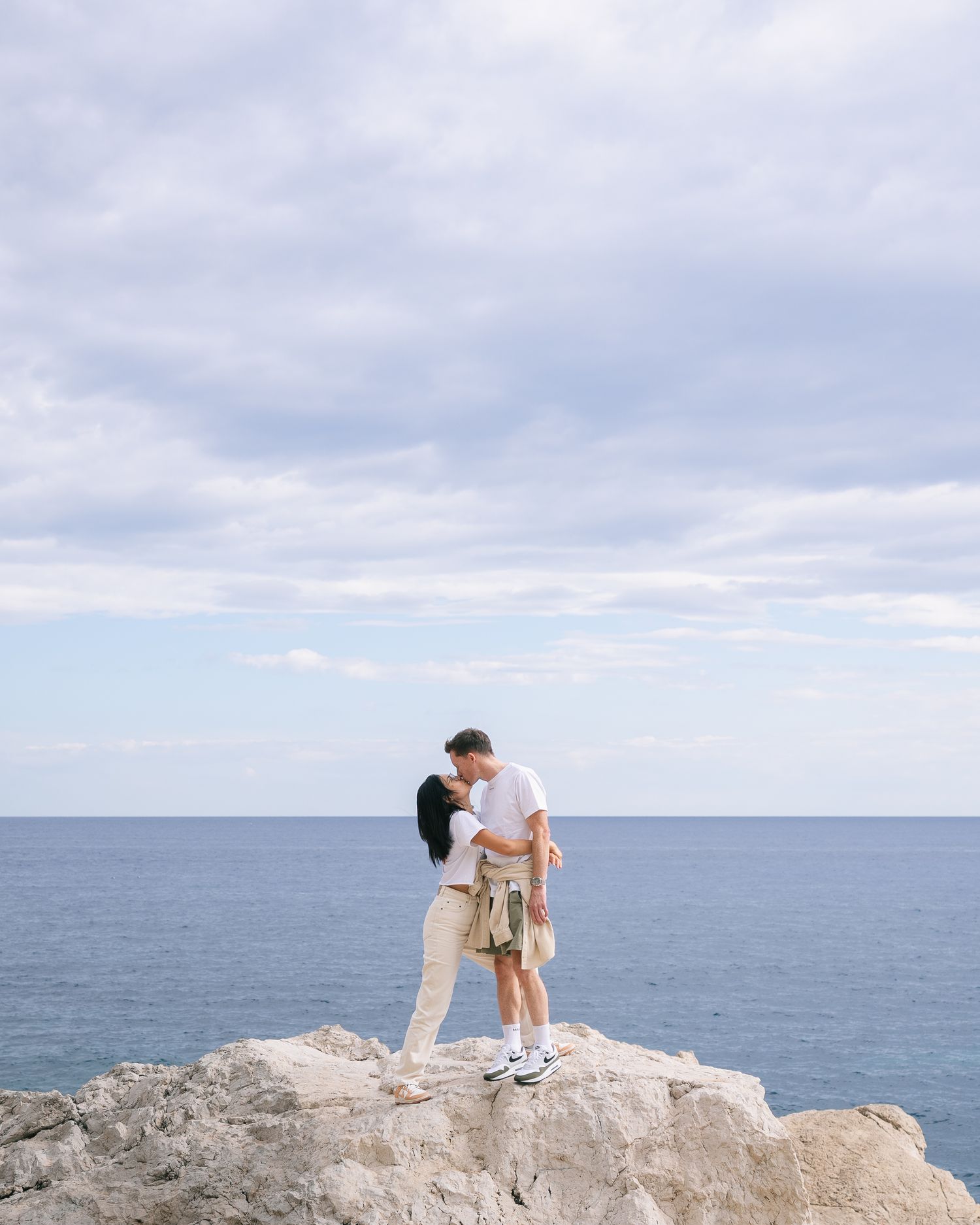 Couple sharing a kiss while standing on rocky cliff overlooking vast blue ocean horizon.