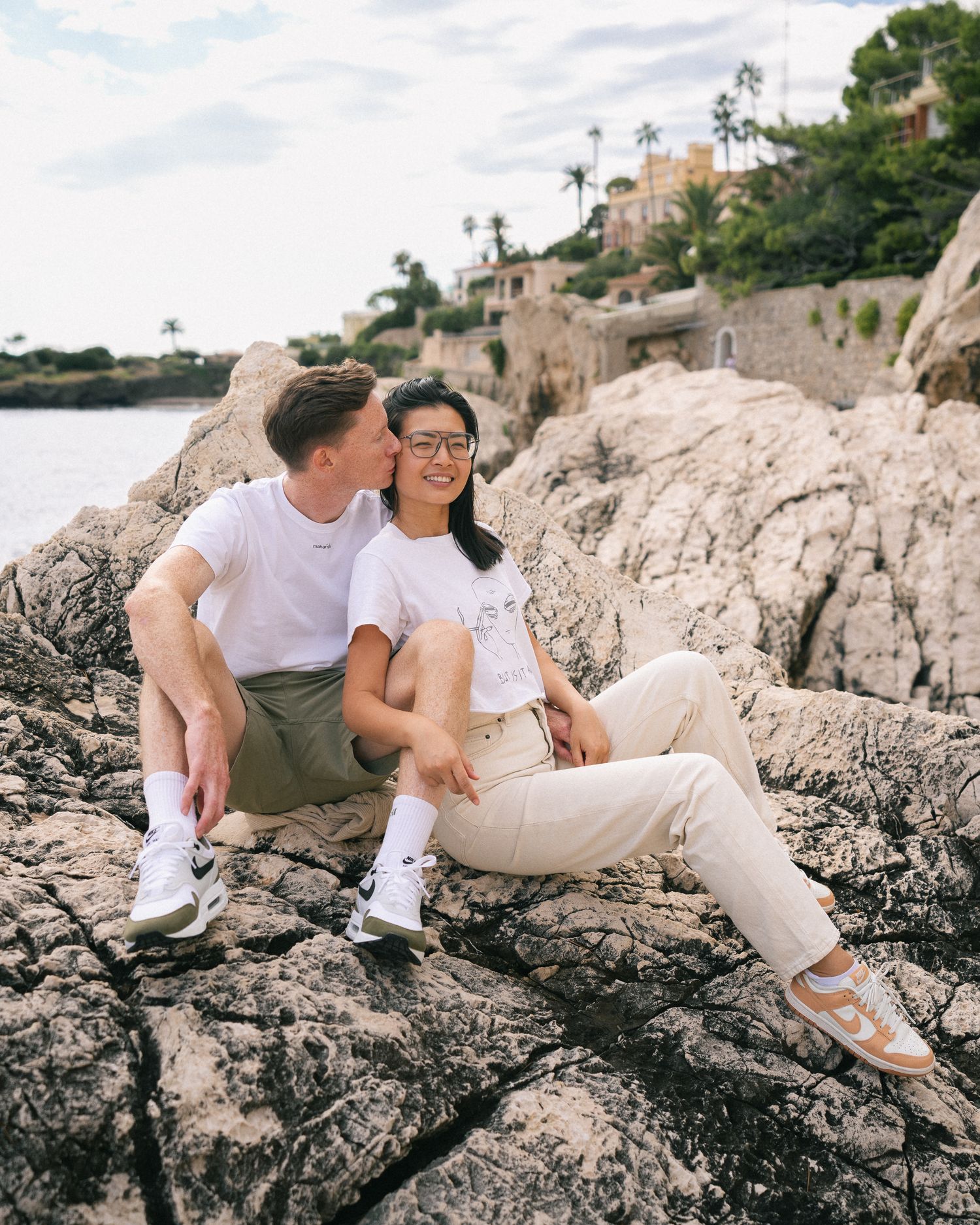 Couple in matching white tees sitting on rocky coastline, man kissing womans cheek at sunset.