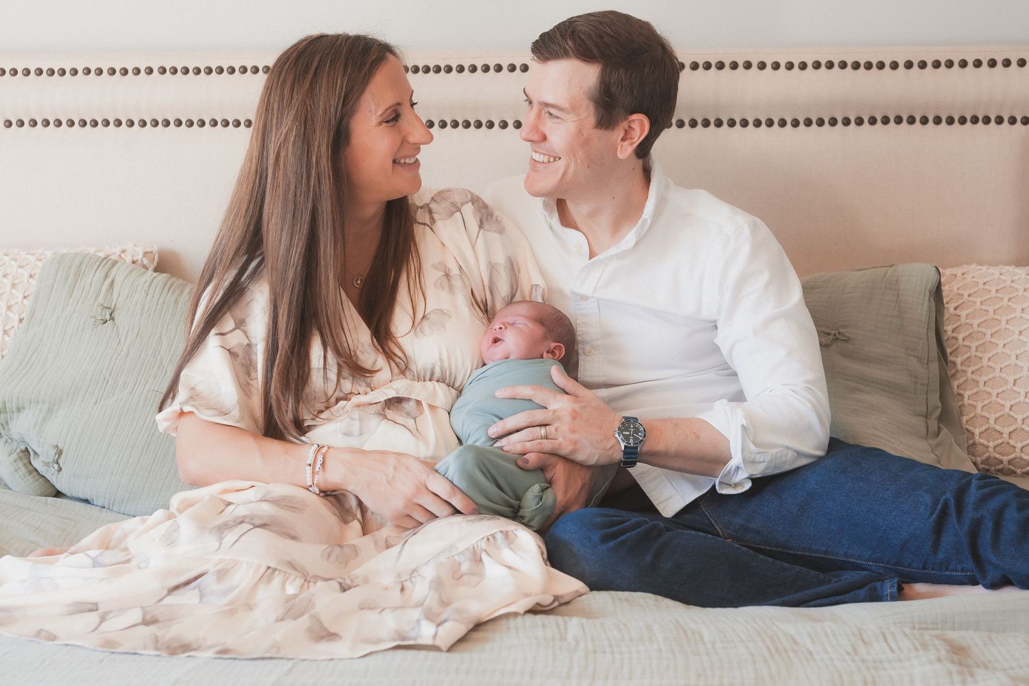 Happy couple sitting on a bed smiling at each other while holding their newborn baby wrapped in a blanket.