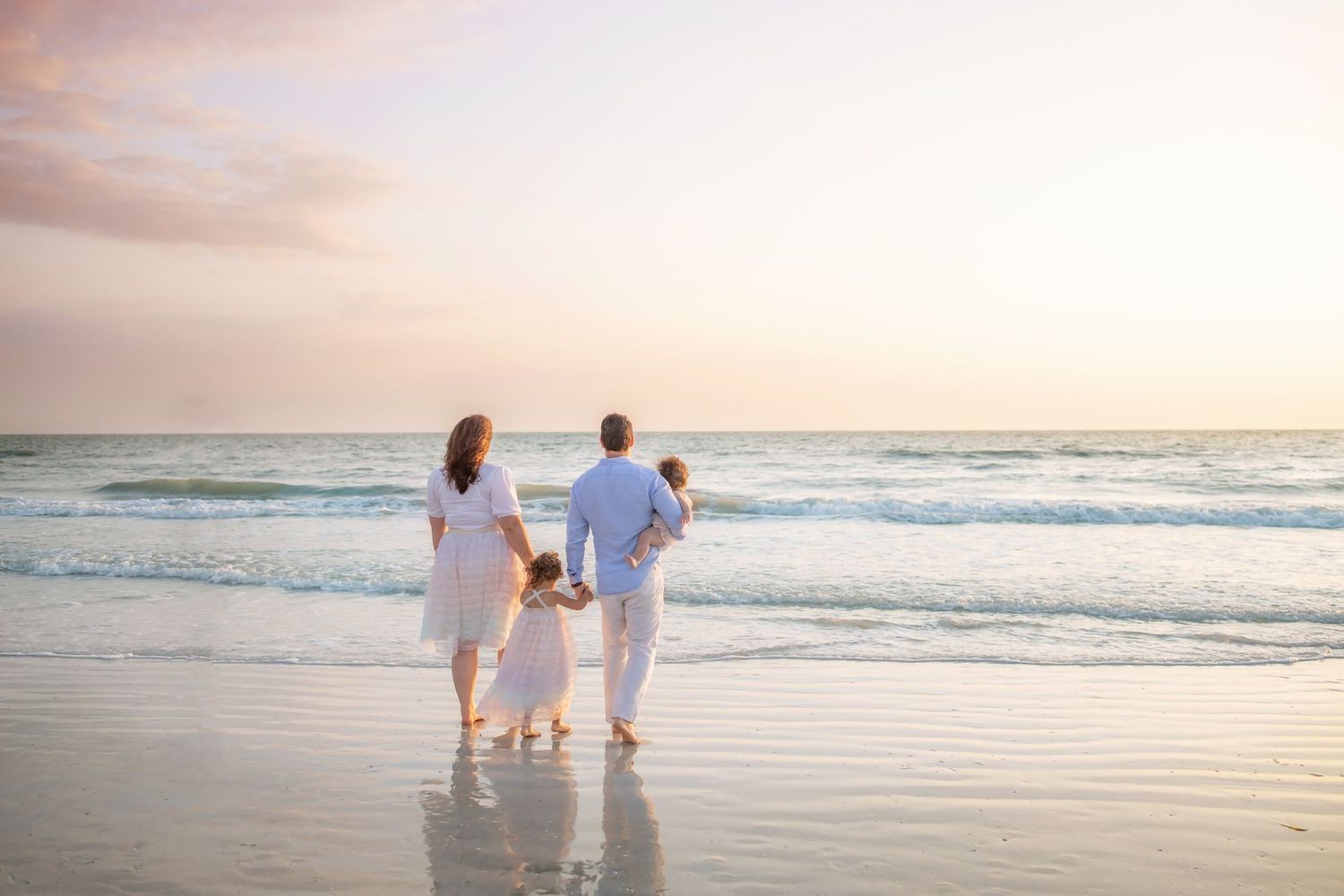 Family of four in white clothing walking on a sandy beach at sunset, holding hands near the ocean waves.