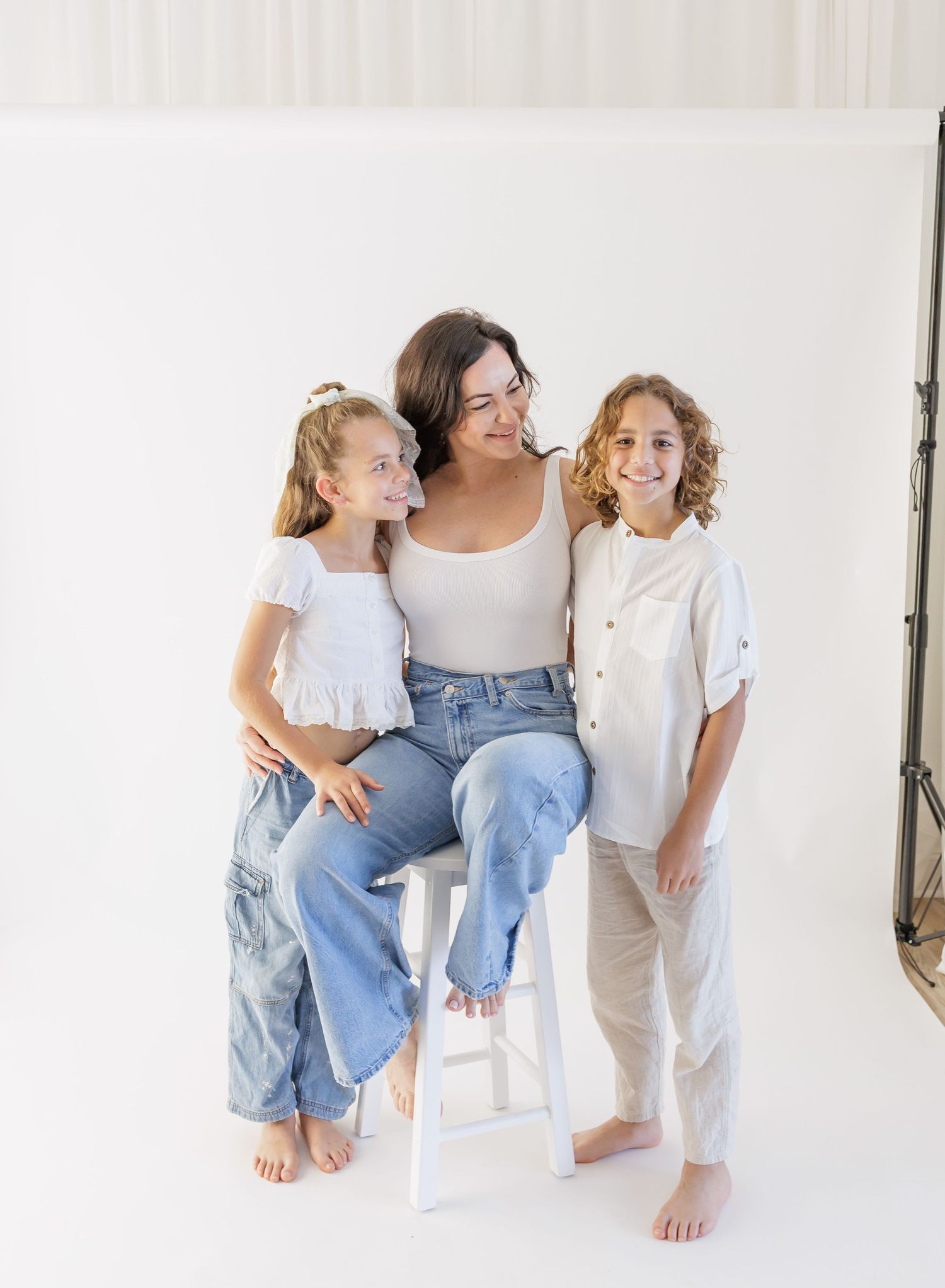 A woman and two children pose together in white outfits against a white backdrop in a bright studio photo.