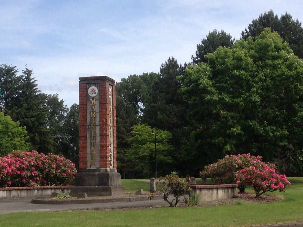 Brick memorial monument surrounded by blooming rhododendrons and tall trees in a lush green park.