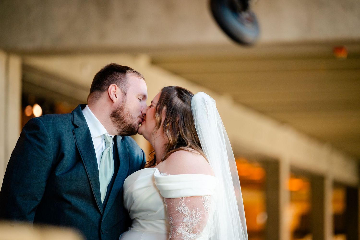 Bride and groom share romantic kiss wearing wedding attire with veil and lace details outdoors.