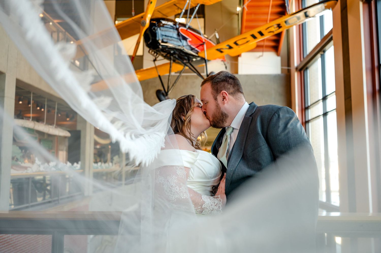 Bride and groom kiss beneath vintage airplane display in rustic industrial wedding venue interior.
