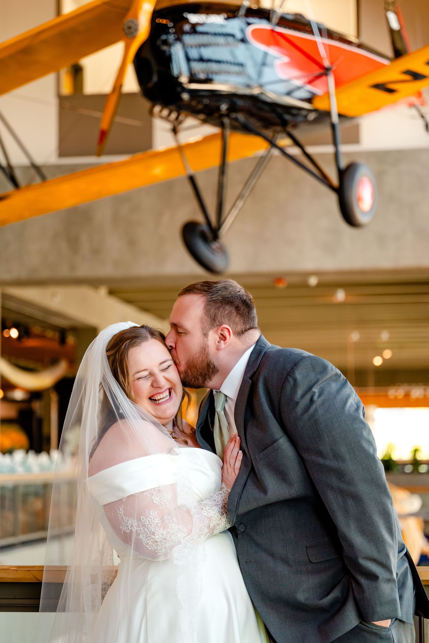 Bride and groom share a joyful moment laughing and kissing beneath a vintage yellow airplane at a museum wedding venue.