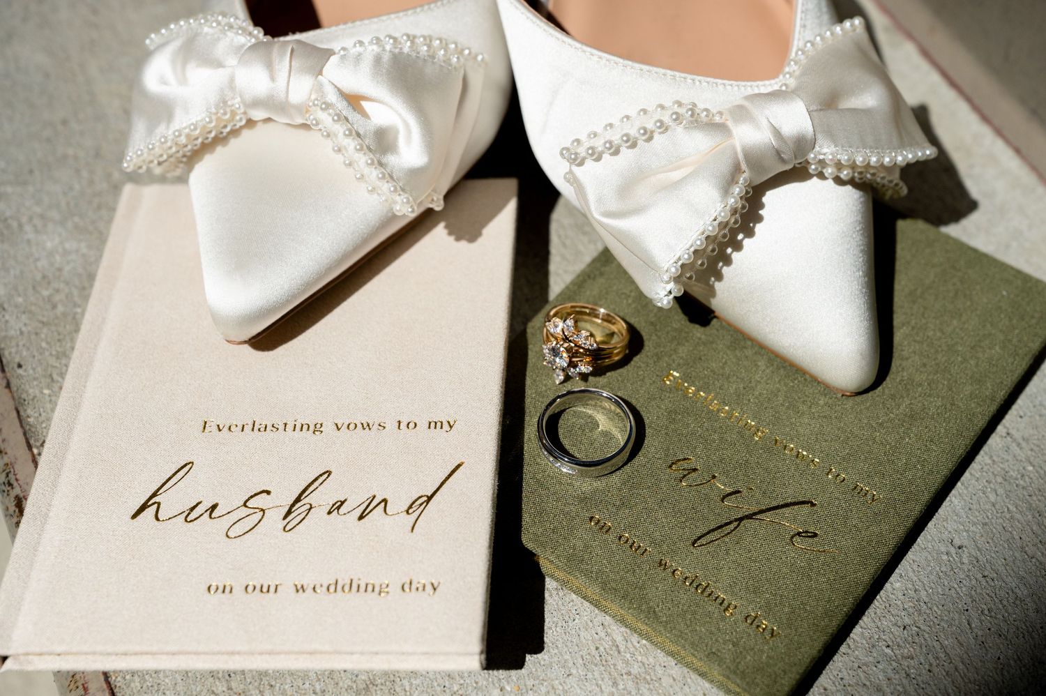 White pearl bow bridal heels beside wedding vows book and rings on a couple's wedding day.
