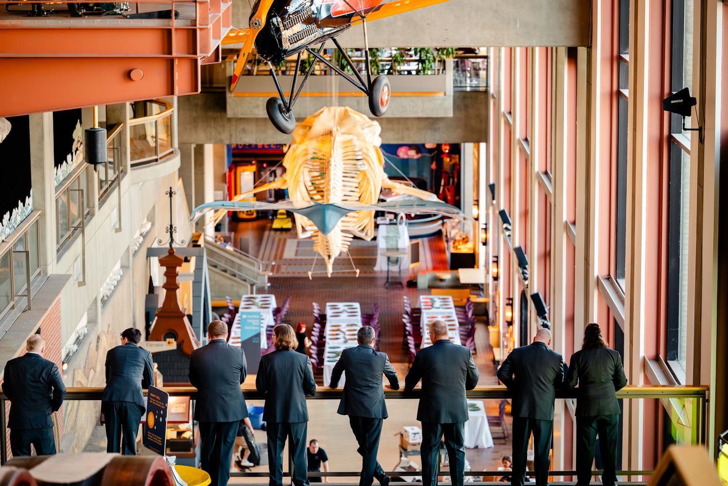 Group of professionals in suits viewing exhibits in a modern museum atrium with hanging aircraft display.