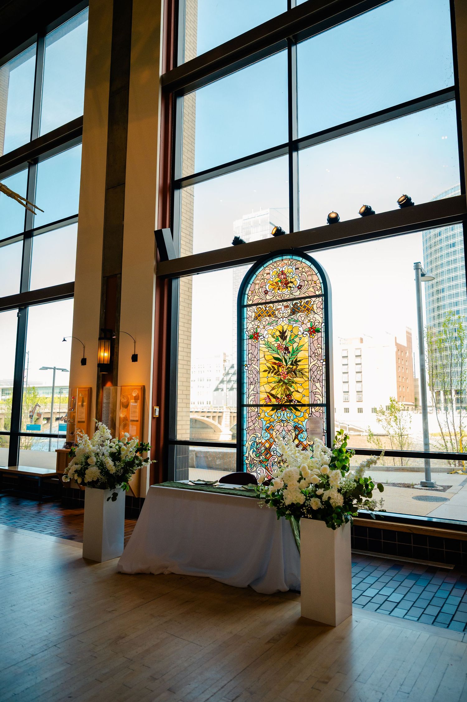Wedding ceremony altar with stained glass and white floral arrangements before large city-view windows.