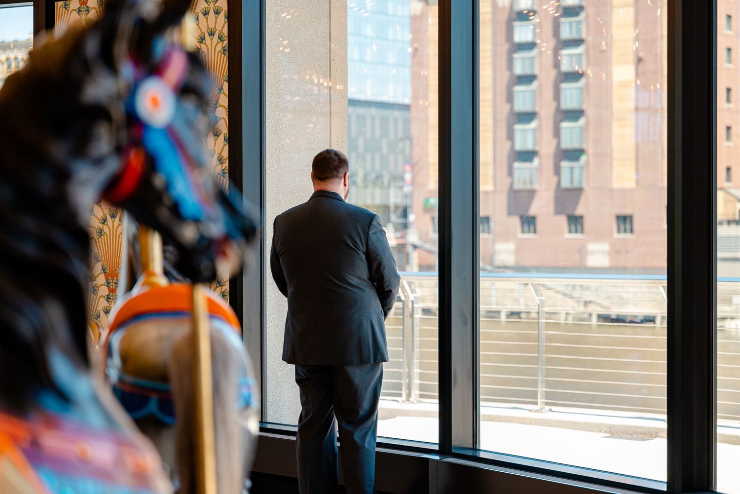 A businessman in a suit stands gazing out large windows at a city skyline, with colorful artwork visible nearby.
