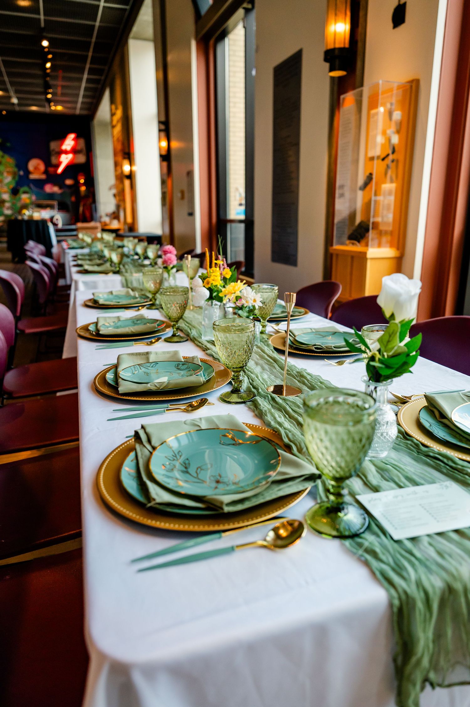 Elegant wedding reception table set with gold chargers, sage green plates, green goblets, and floral centerpieces on white linen.