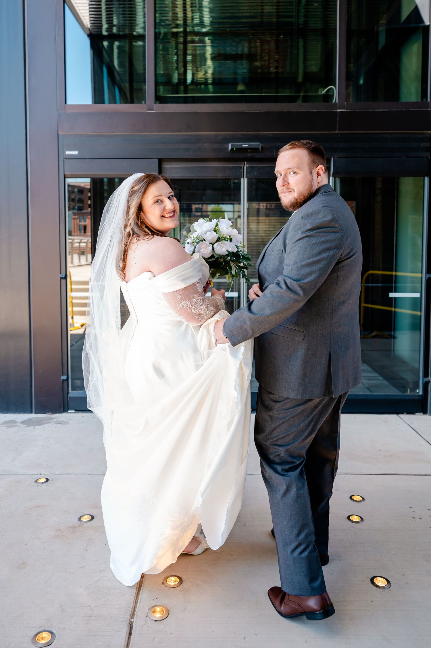 Bride in white gown with veil and groom in gray suit pose together outside modern building on their wedding day.