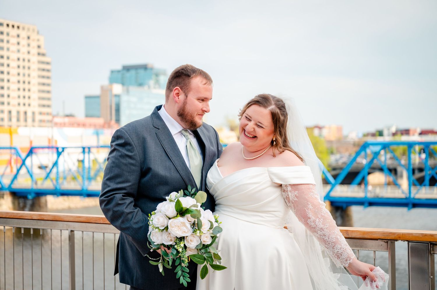 Bride and groom laughing together on a riverside bridge, she holds white roses bouquet, city skyline and blue bridge behind them.