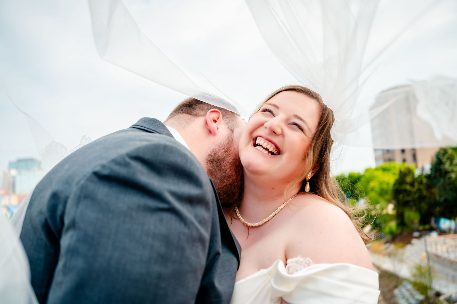 A joyful bride laughs as her groom leans in to kiss her cheek during an outdoor wedding celebration.