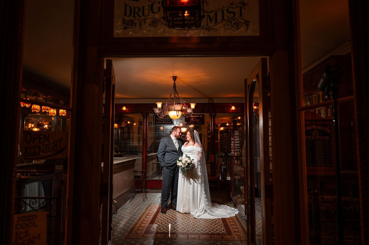 Bride and groom share a romantic kiss in a warmly lit vintage drugstore doorway on their wedding day.