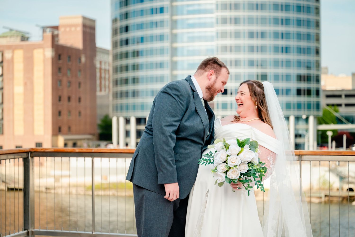 Bride and groom laughing together on a bridge with city skyline backdrop, bride holding white bouquet.