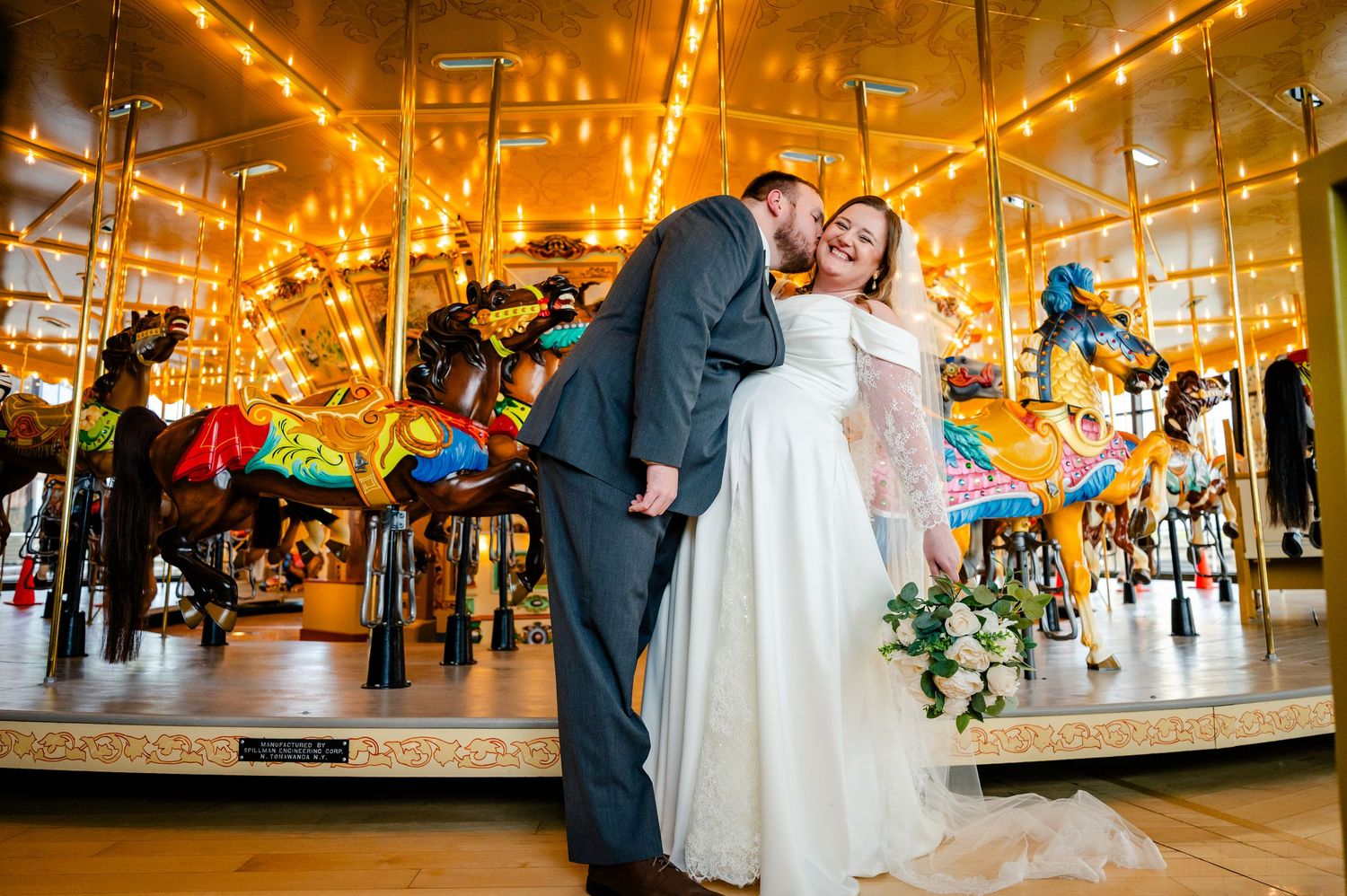 Bride and groom share a kiss in front of a glowing carousel at their wedding reception.