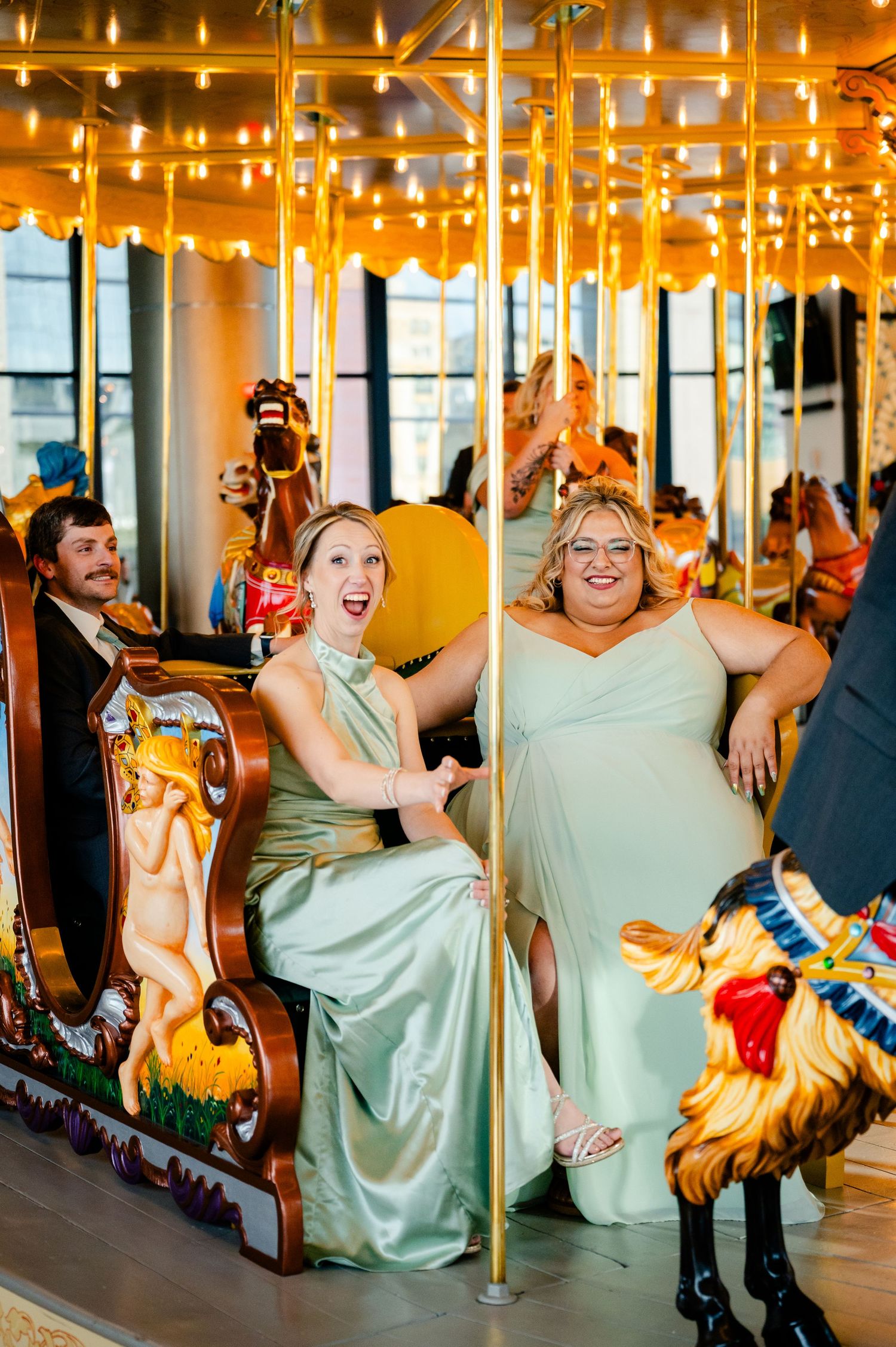 Two bridesmaids in sage green gowns laugh joyfully while riding a ornate golden carousel at a wedding reception.
