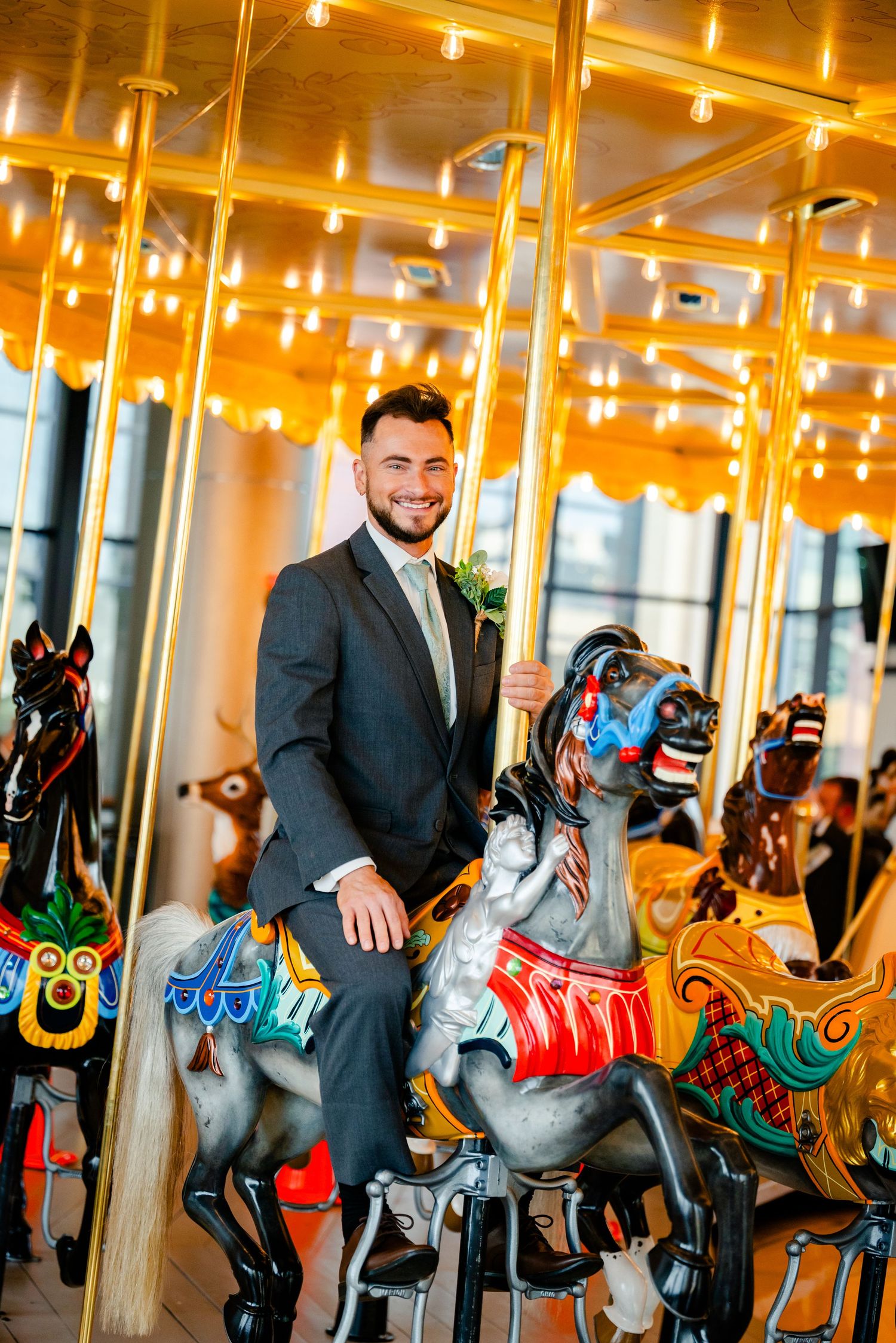 Smiling groom in dark suit sitting on colorful carousel horse surrounded by festive lights.