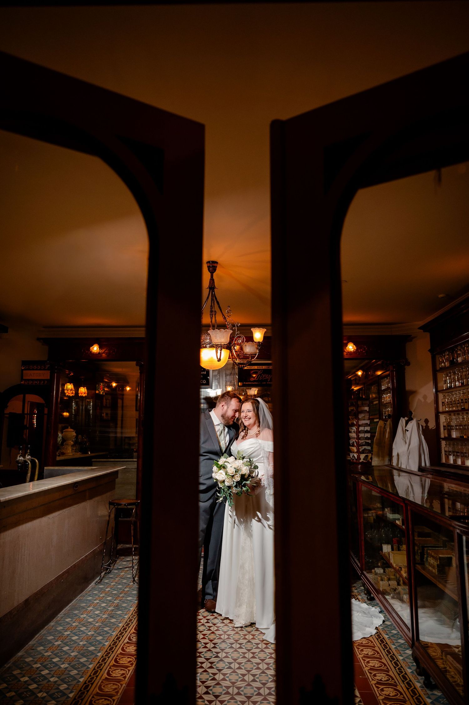 Bride and groom share a romantic moment framed by wooden doorway in a warmly lit vintage interior.