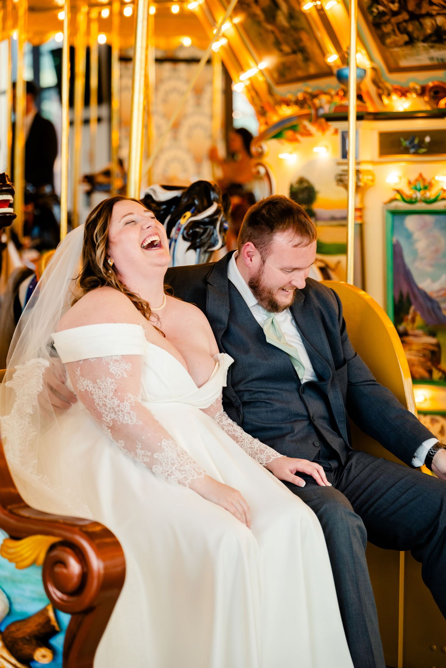 A laughing bride and groom in wedding attire enjoy a romantic carousel ride together, surrounded by warm golden lights.