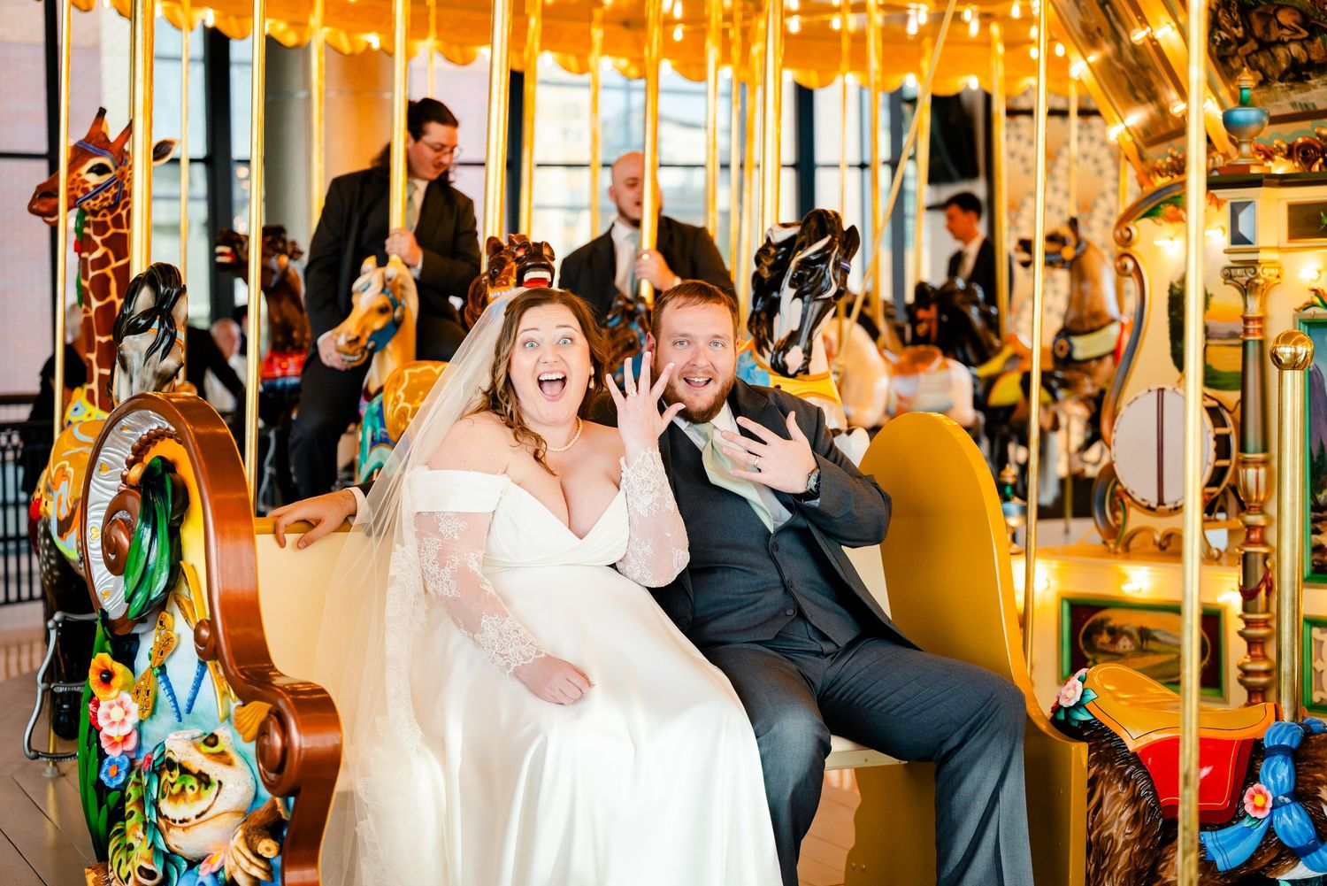 Joyful bride and groom riding a carousel, laughing and showing off wedding rings at their reception.