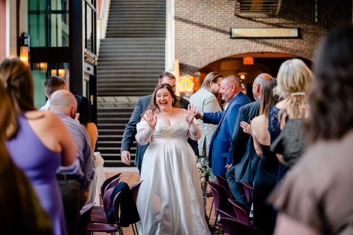 Bride walks down the aisle smiling as wedding guests applaud at indoor ceremony venue.