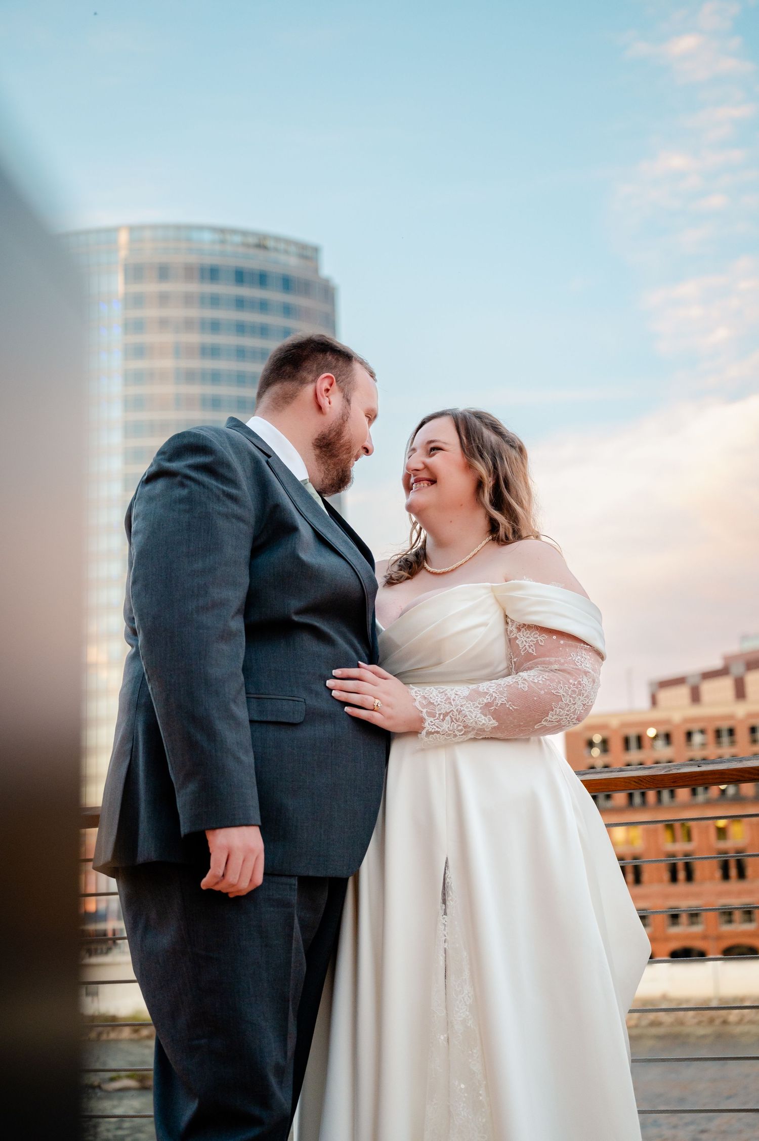 A bride and groom share a romantic moment on a rooftop with a city skyline backdrop at sunset.