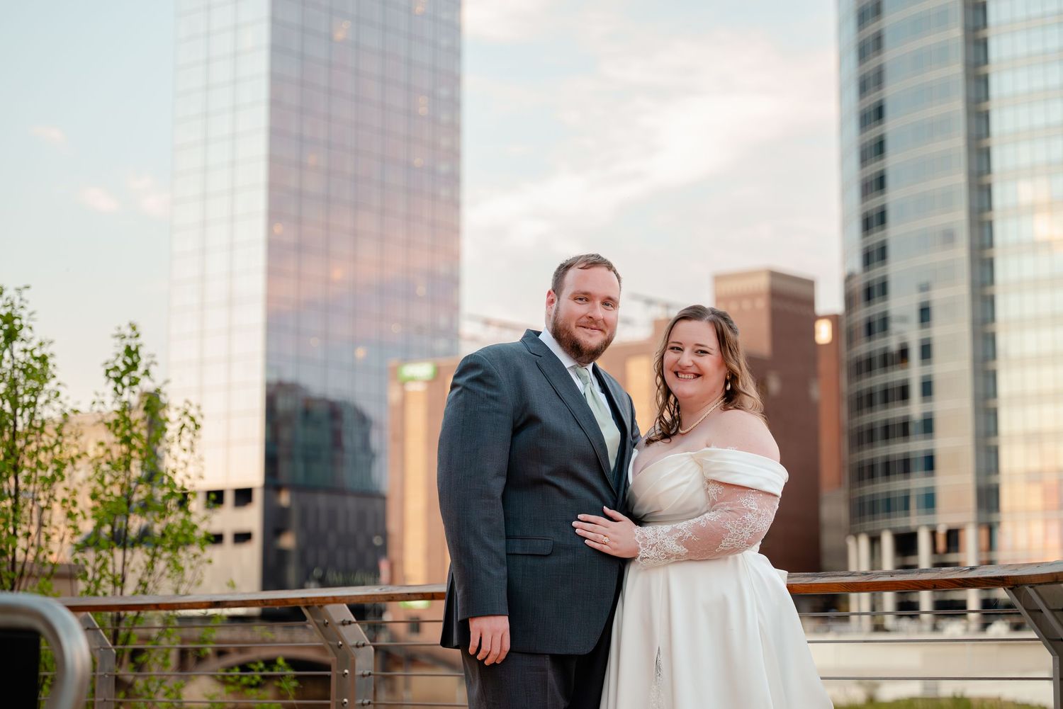 Smiling bride and groom pose together on a rooftop terrace with city skyscrapers at sunset.