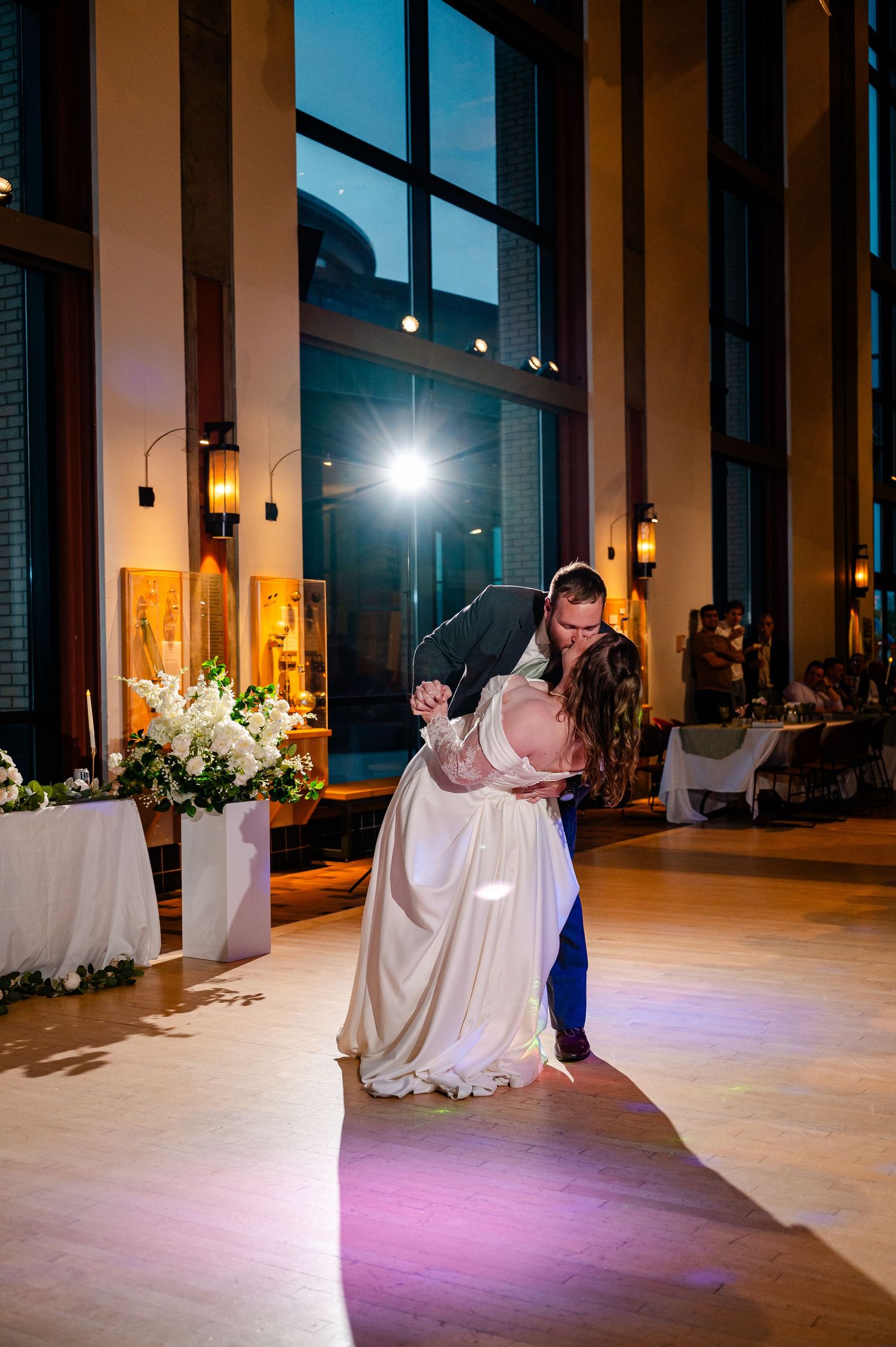 Bride and groom share romantic first dance dip in elegant wedding venue with large windows and floral arrangements.
