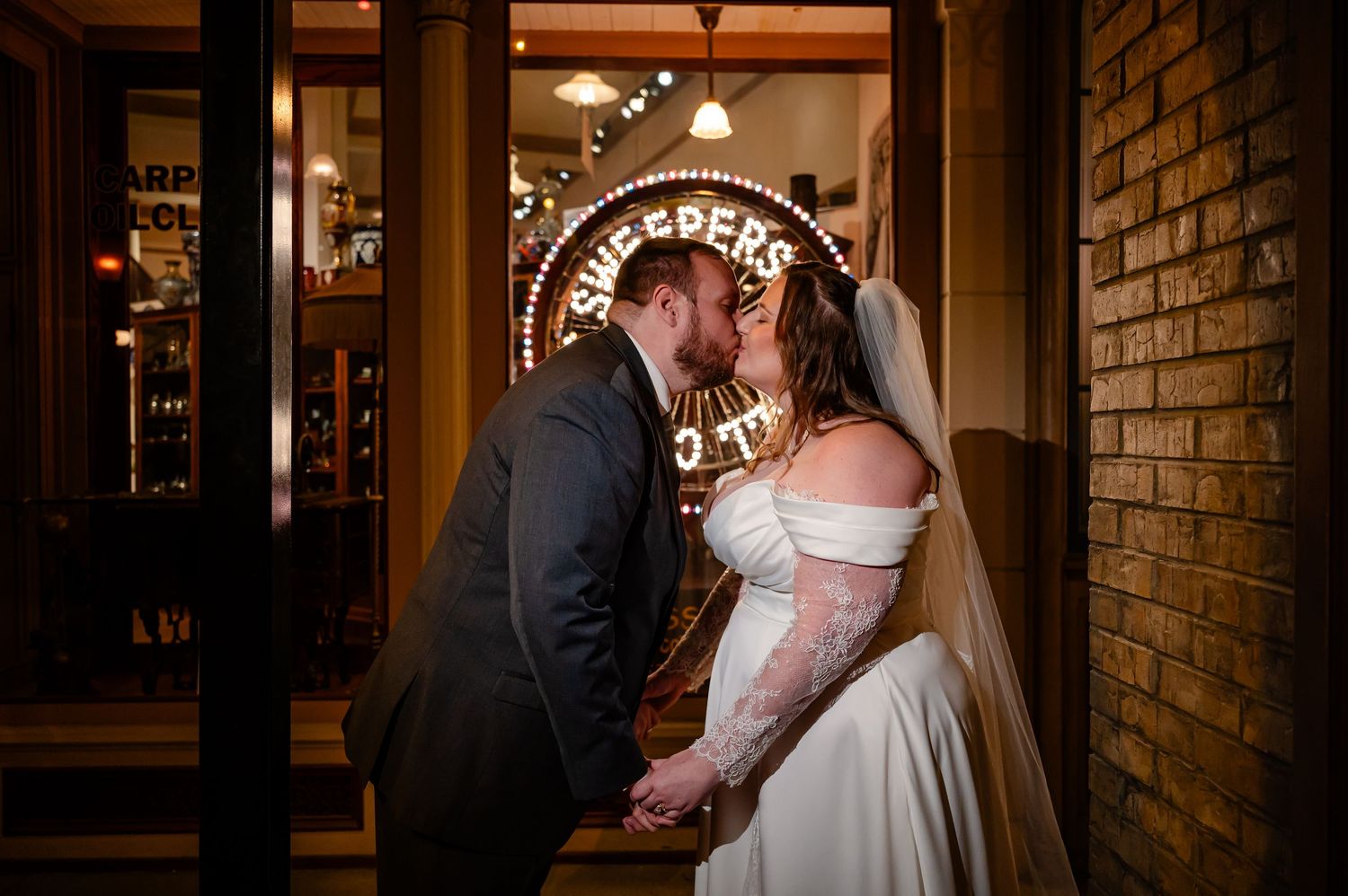 Bride and groom share a romantic kiss outside a warmly lit venue at night, bride wearing lace gown and veil.