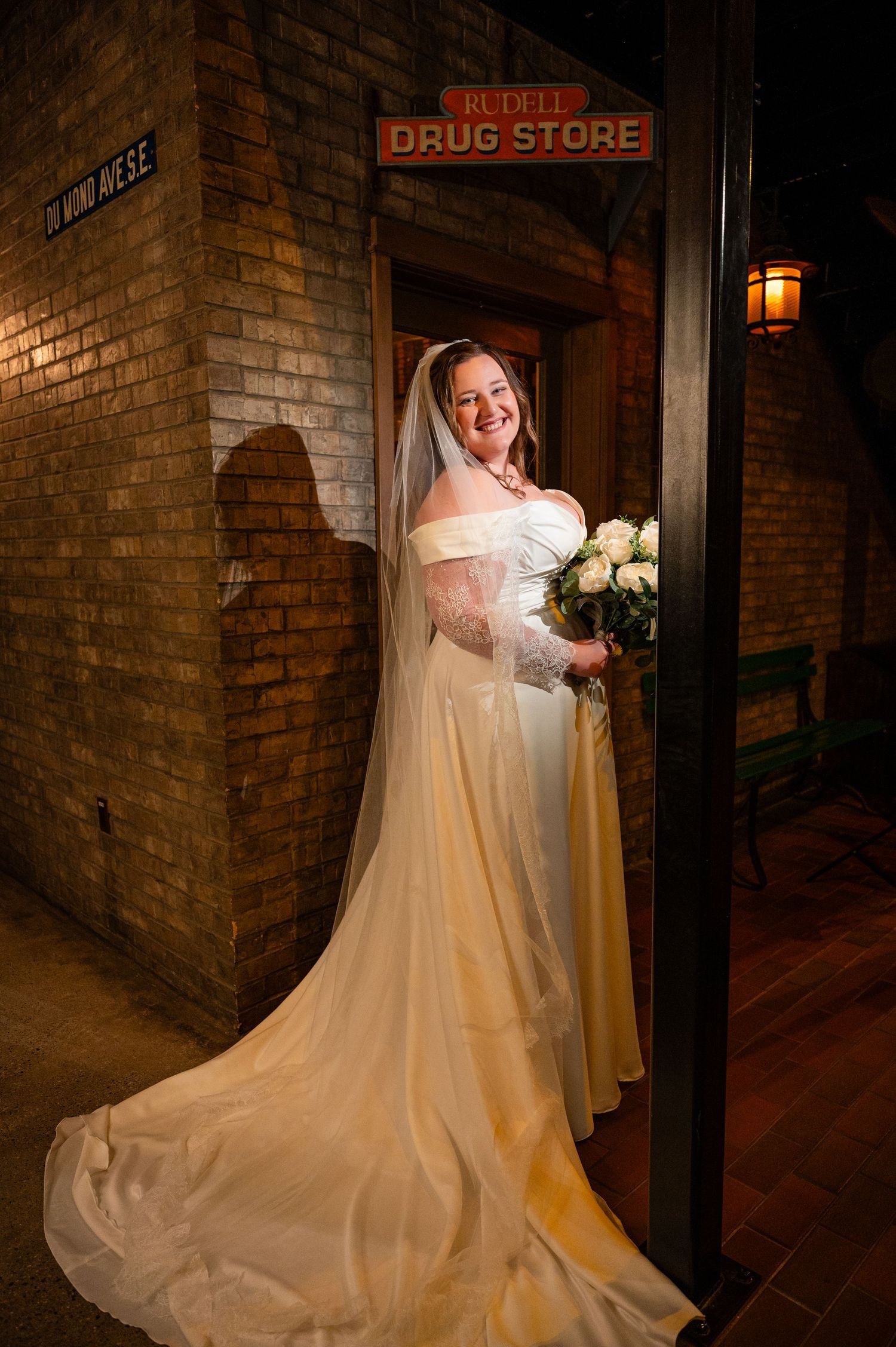 Bride in white gown and veil holding bouquet, posing by Rudell Drug Store sign on brick wall.