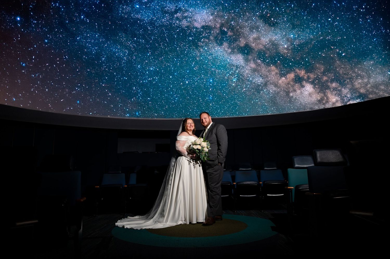 Bride and groom pose under a stunning Milky Way galaxy projection at a planetarium wedding venue.