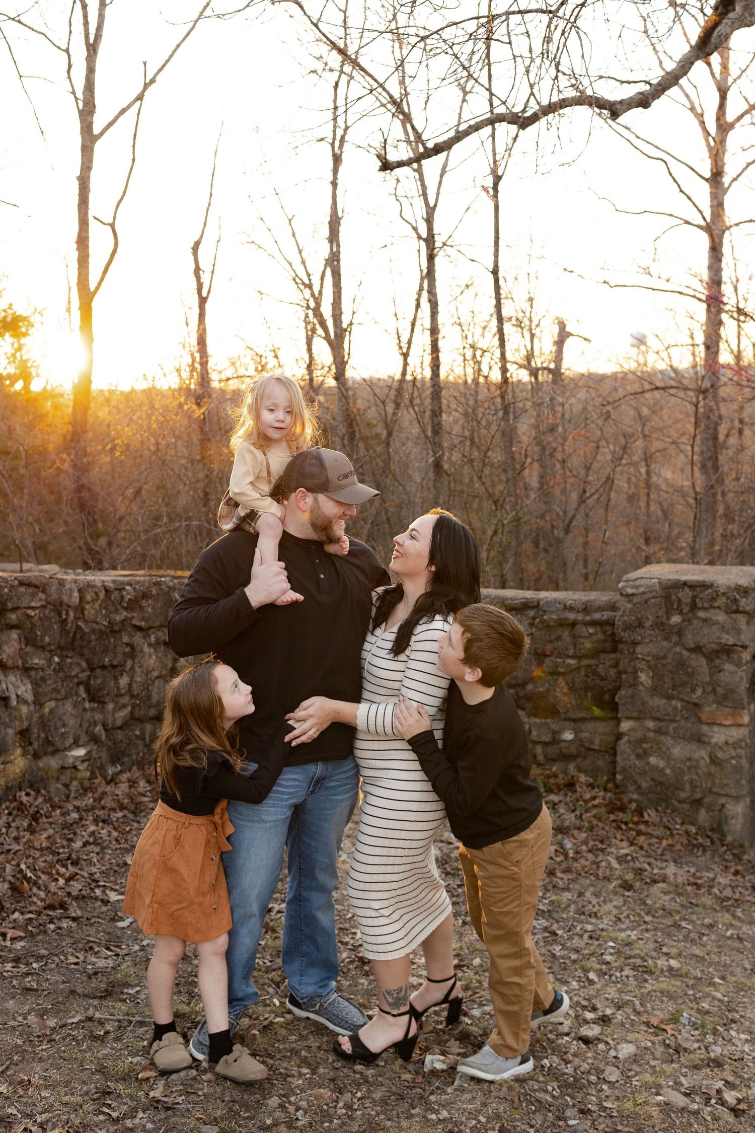 Family of five posing at golden hour outdoors, parents with three young children near a stone wall surrounded by autumn trees.