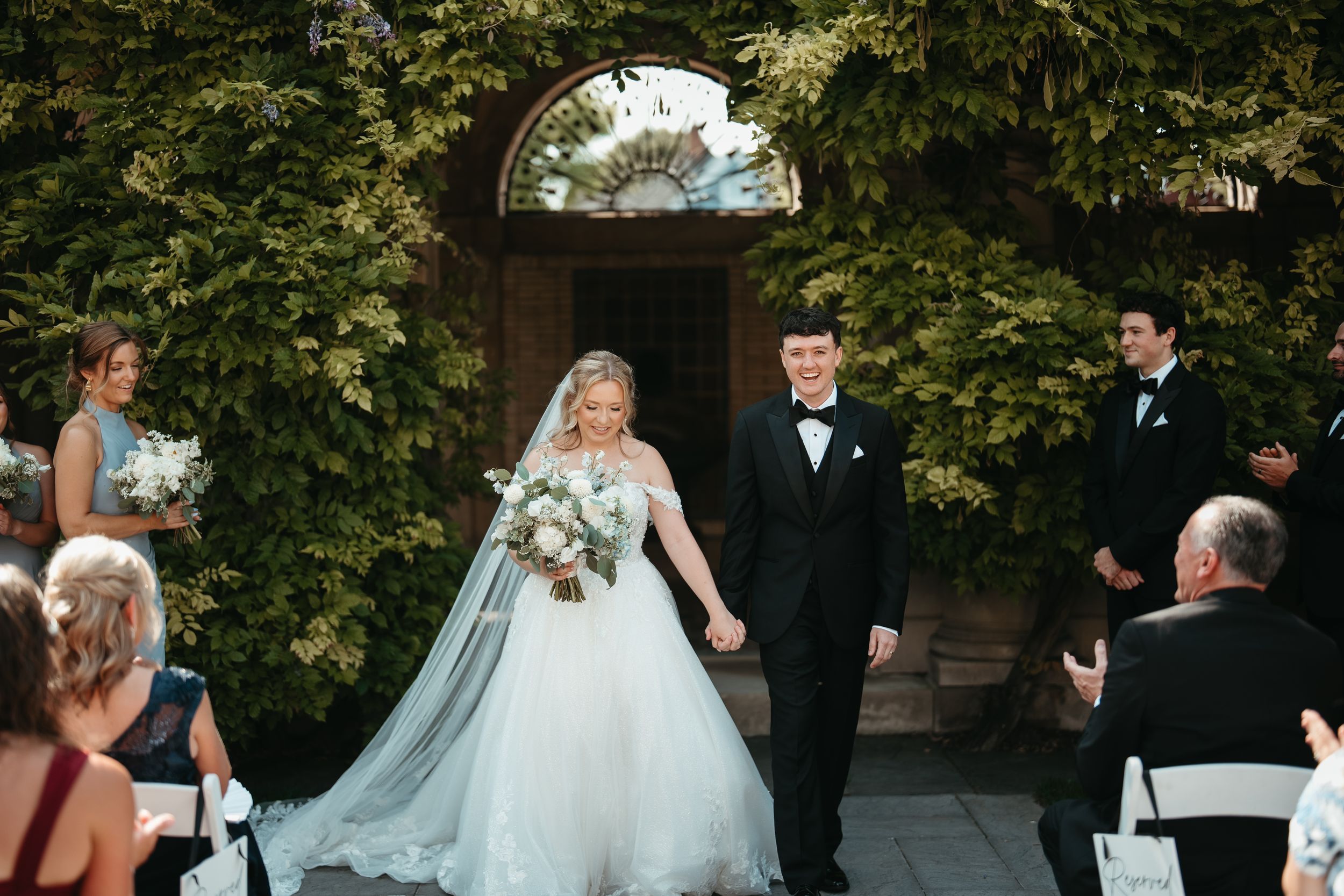 Bride and groom walk hand-in-hand down outdoor aisle after wedding ceremony, surrounded by lush greenery and seated guests.