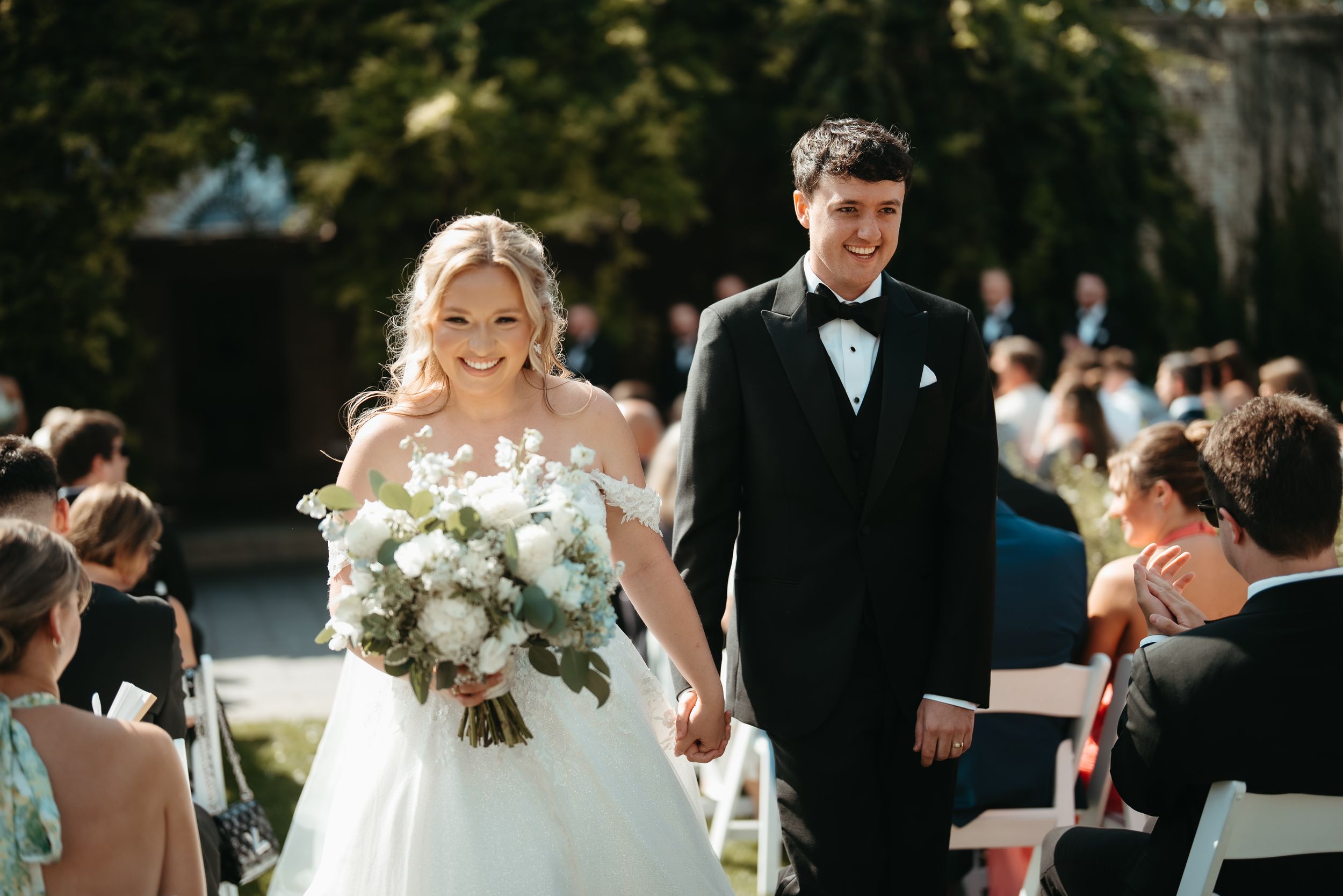 Bride and groom walk down the aisle hand in hand at an outdoor wedding ceremony, bride holding white floral bouquet.