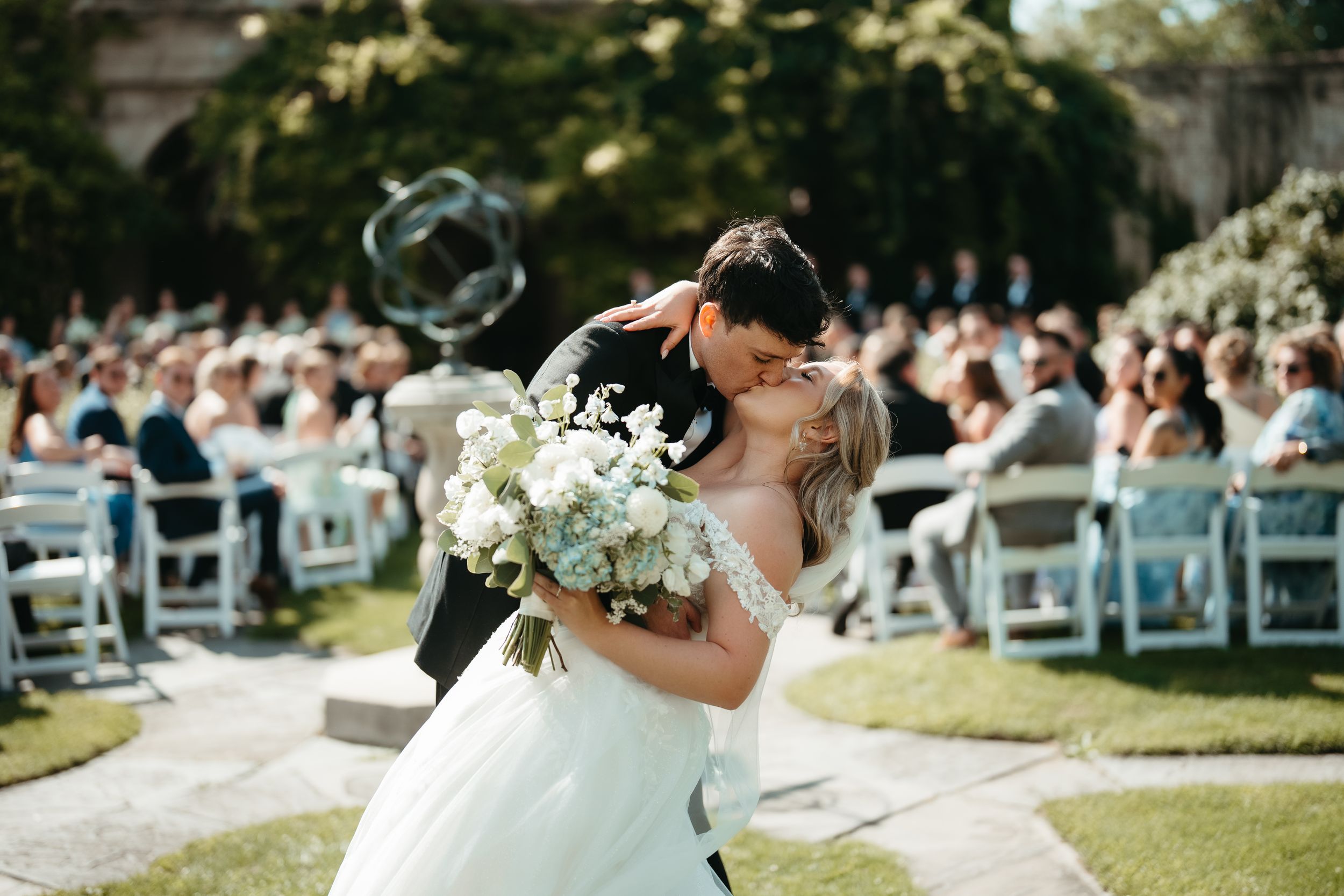 Bride and groom share first kiss at outdoor wedding ceremony with white chairs and guests in background.