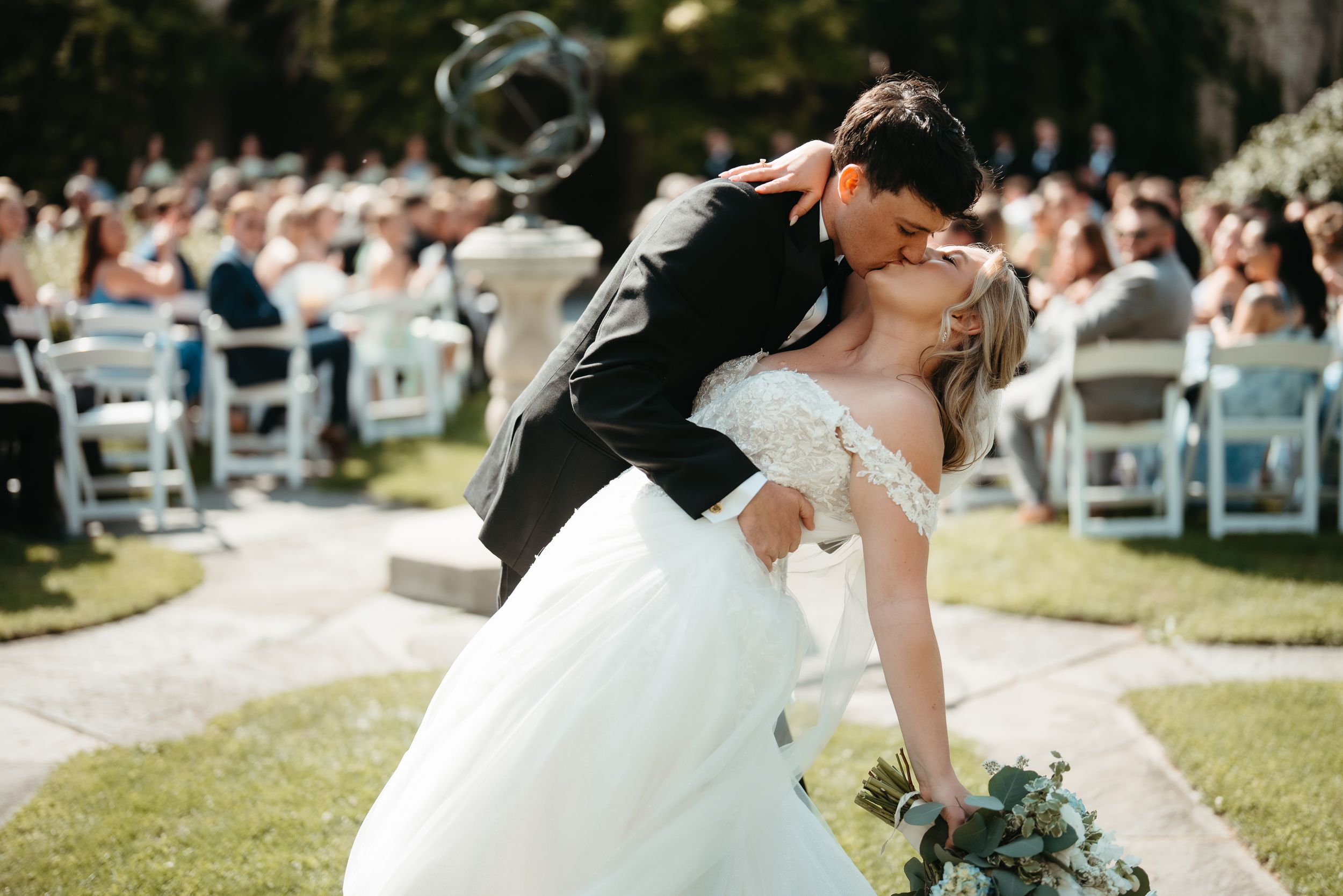 Bride and groom share romantic dip kiss at outdoor wedding ceremony with guests seated on white chairs.