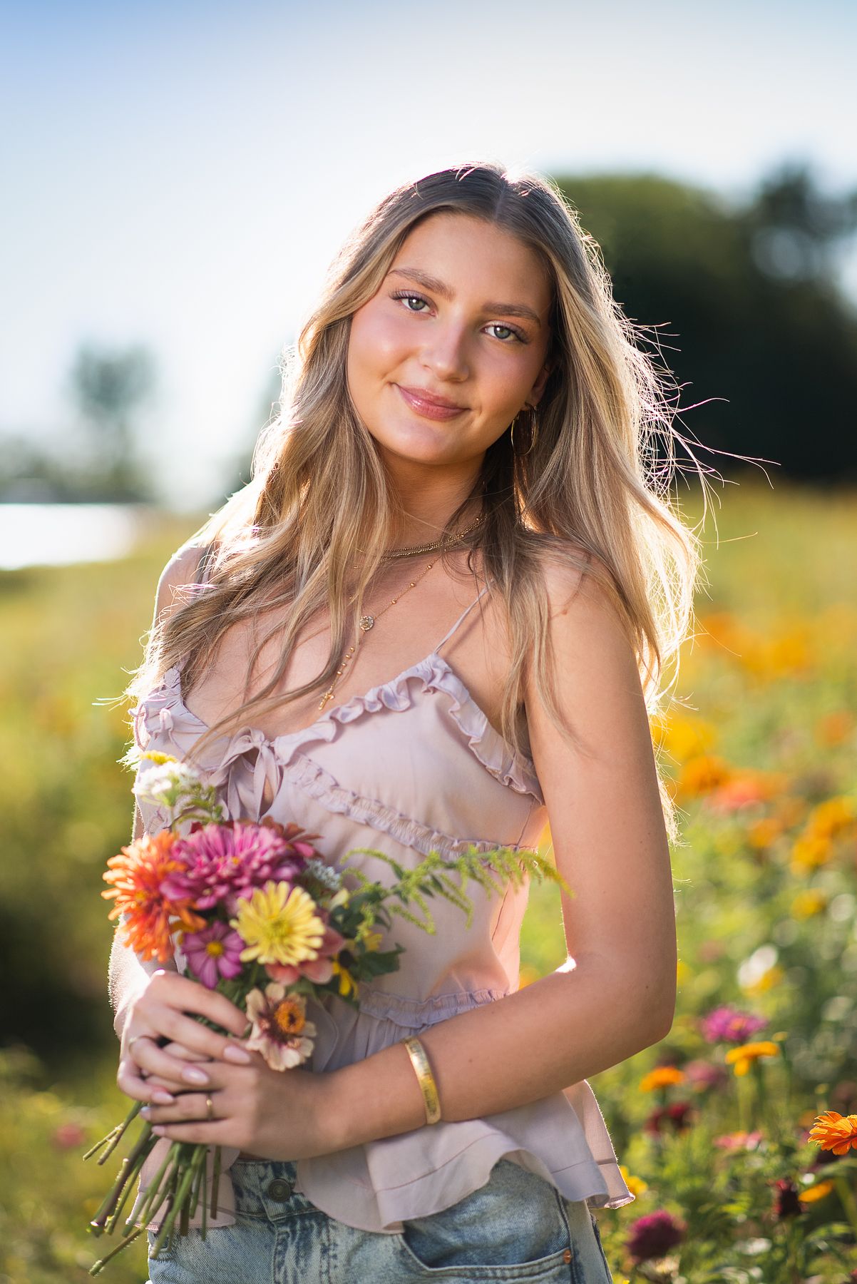 Young blonde woman holding colorful flower bouquet in sunny flower field, wearing pink top and jeans.