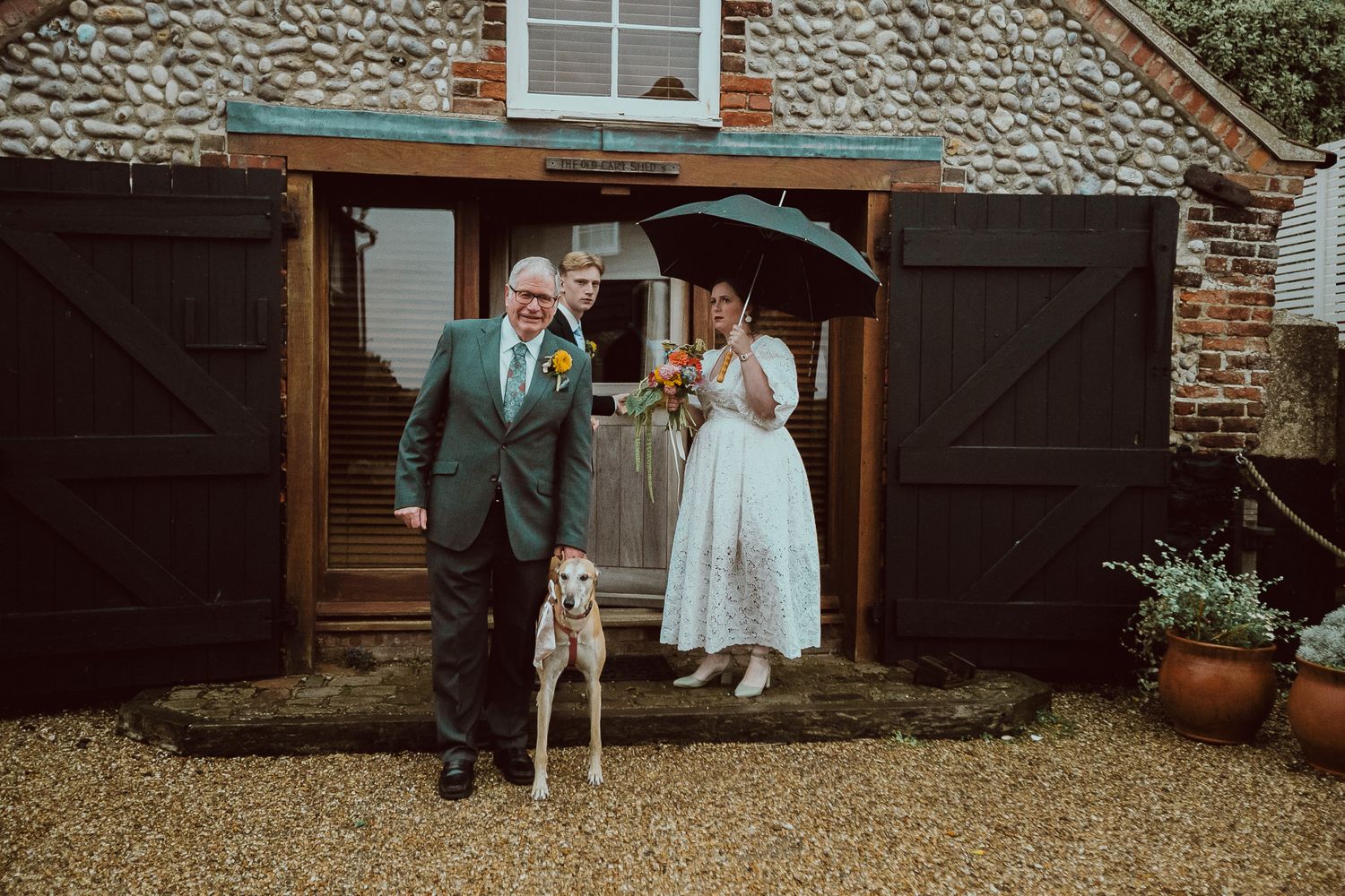 Bride and groom stand with a white dog outside rustic barn doors, bride holding umbrella on a rainy wedding day.